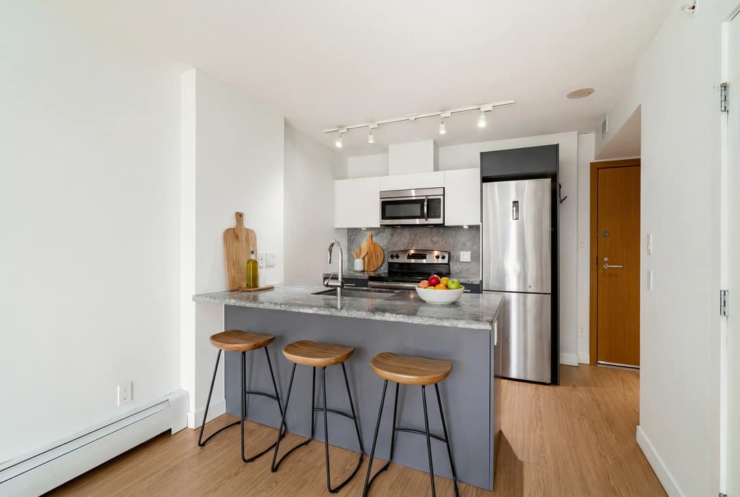 Before and after virtual staging of a Downtown Vancouver condo kitchen, adding wood bar stools, a cutting board, and fruit bowl to the granite island to bring warmth to the stainless steel and slate finishes.