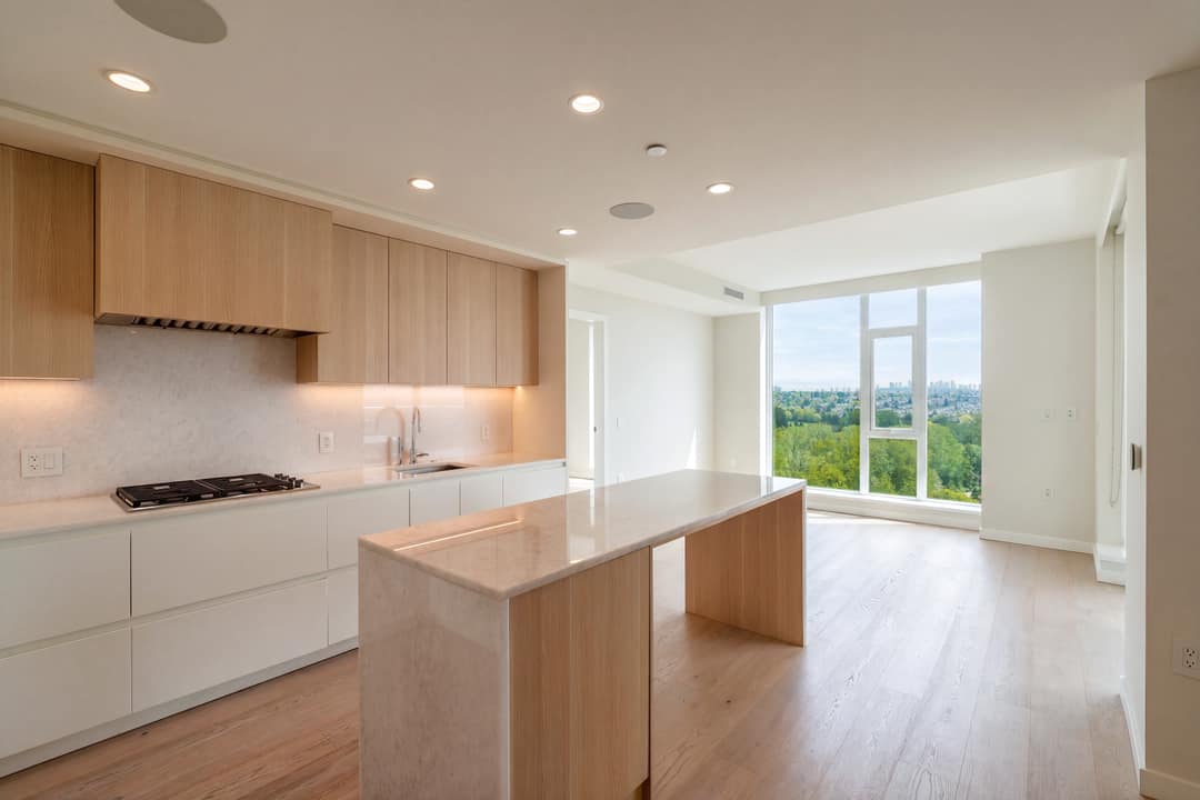 Bright empty kitchen in a Cambie Gardens condo featuring wood cabinetry, a waterfall island, gas cooktop, and floor-to-ceiling windows overlooking green treetops and the city skyline.