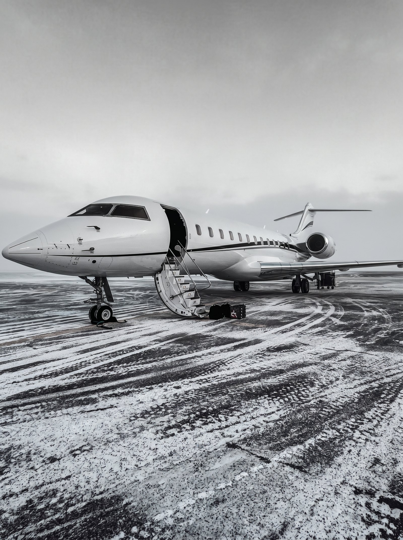 Black-and-white photograph of a midsize business jet being de-iced on a snowy Arctic apron at dusk, ground crew in cold-