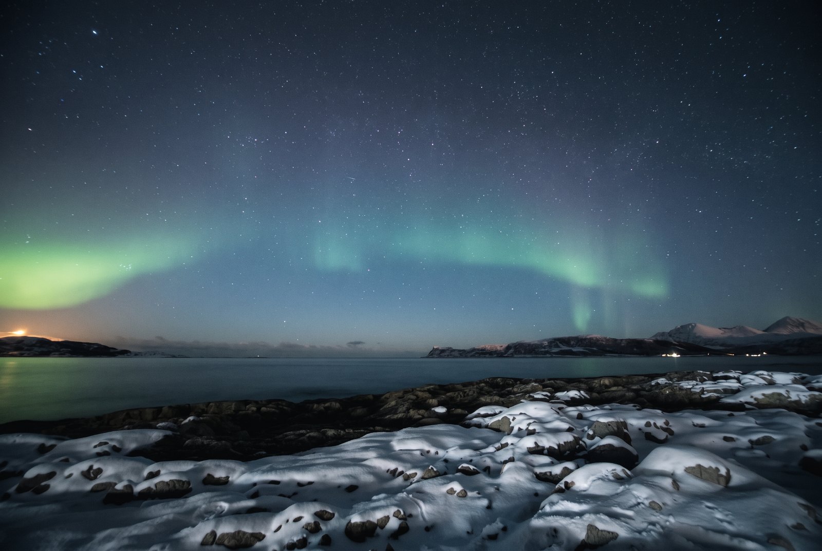 Black-and-white view of Tromsø during polar night: mountains, fjord and city lights under a dark sky with a faint band o