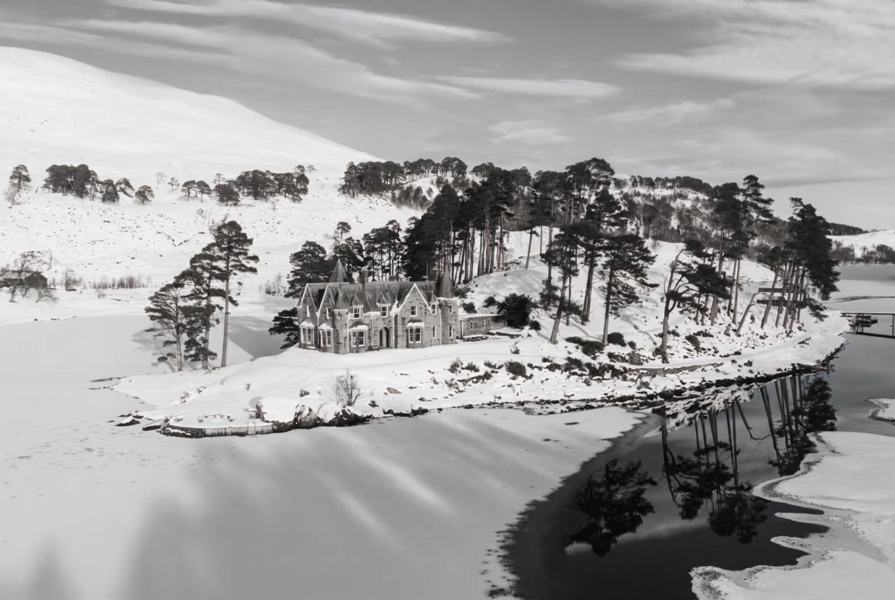 Black-and-white photograph of a light helicopter set down on the lawn or parkland of a country estate, a stone lodge or