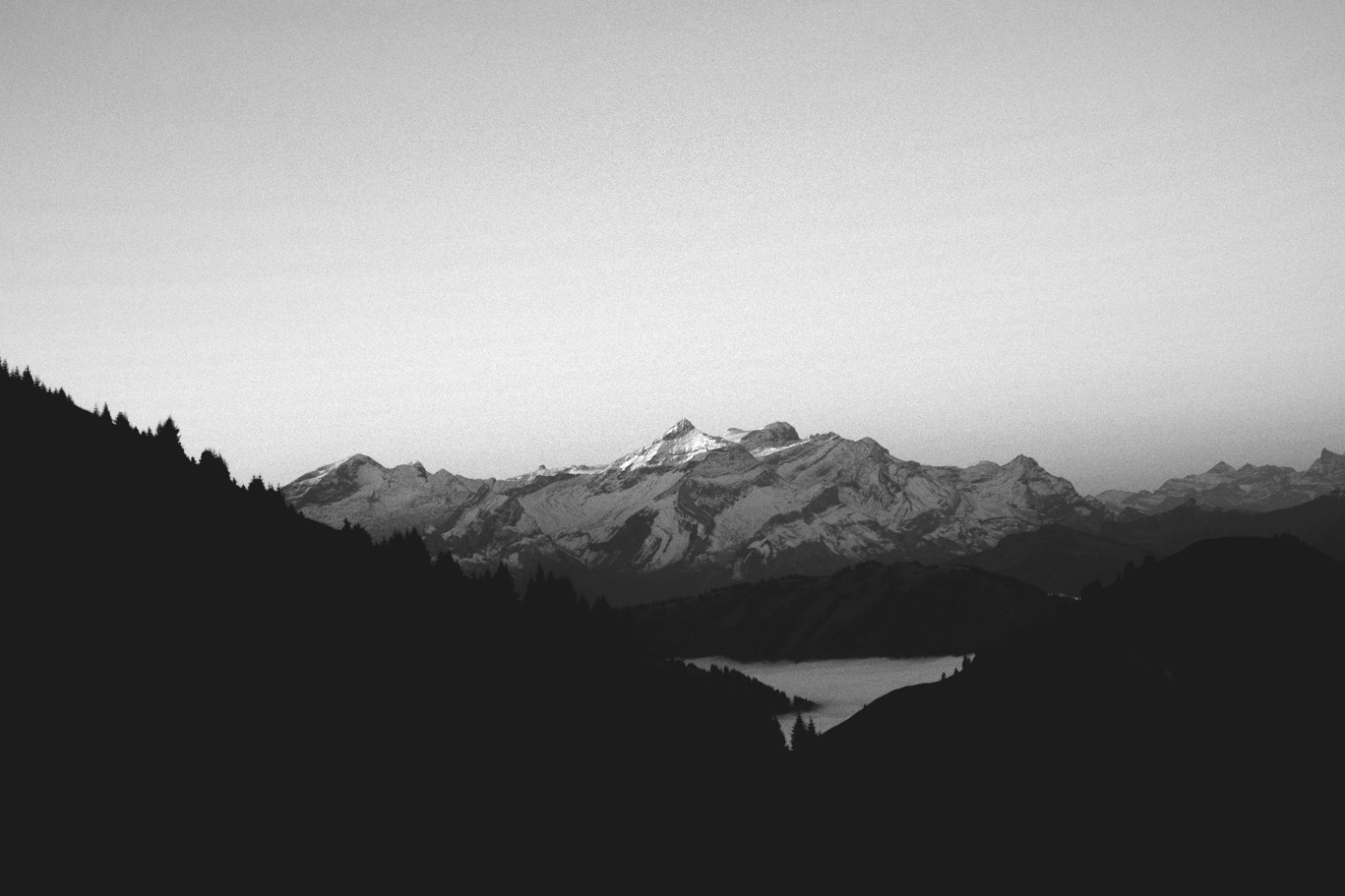 Black-and-white Alpine panorama at dusk, with snow-capped peaks above dark forested slopes and a valley.