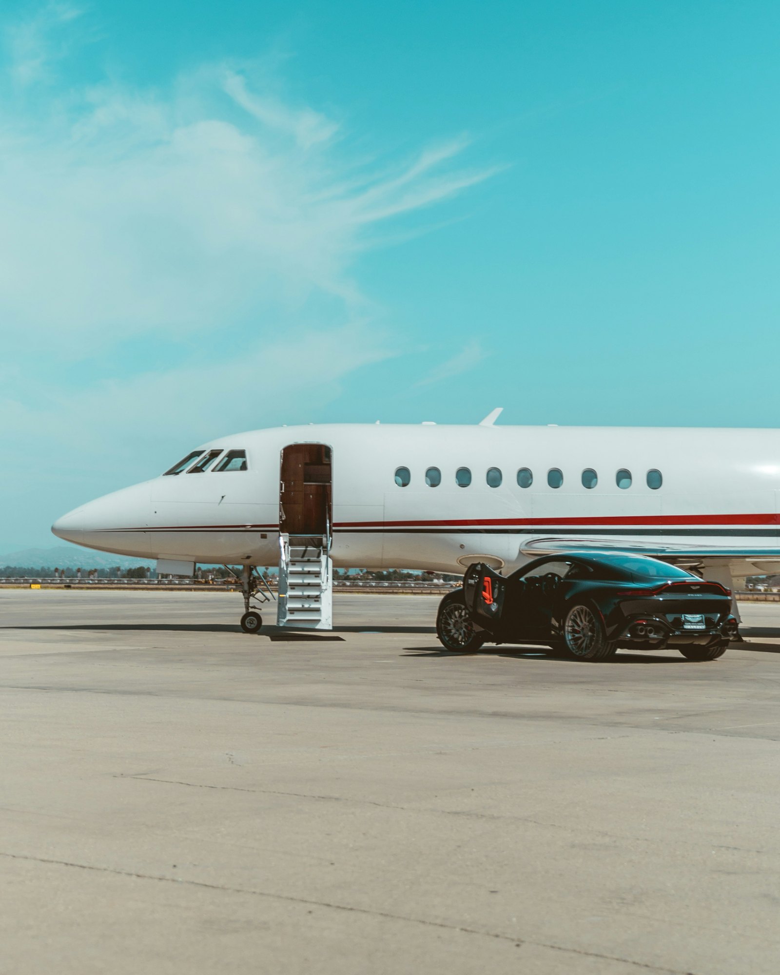 A white business jet parked on a sunlit apron under a blue sky, with a dark car beside its airstair.