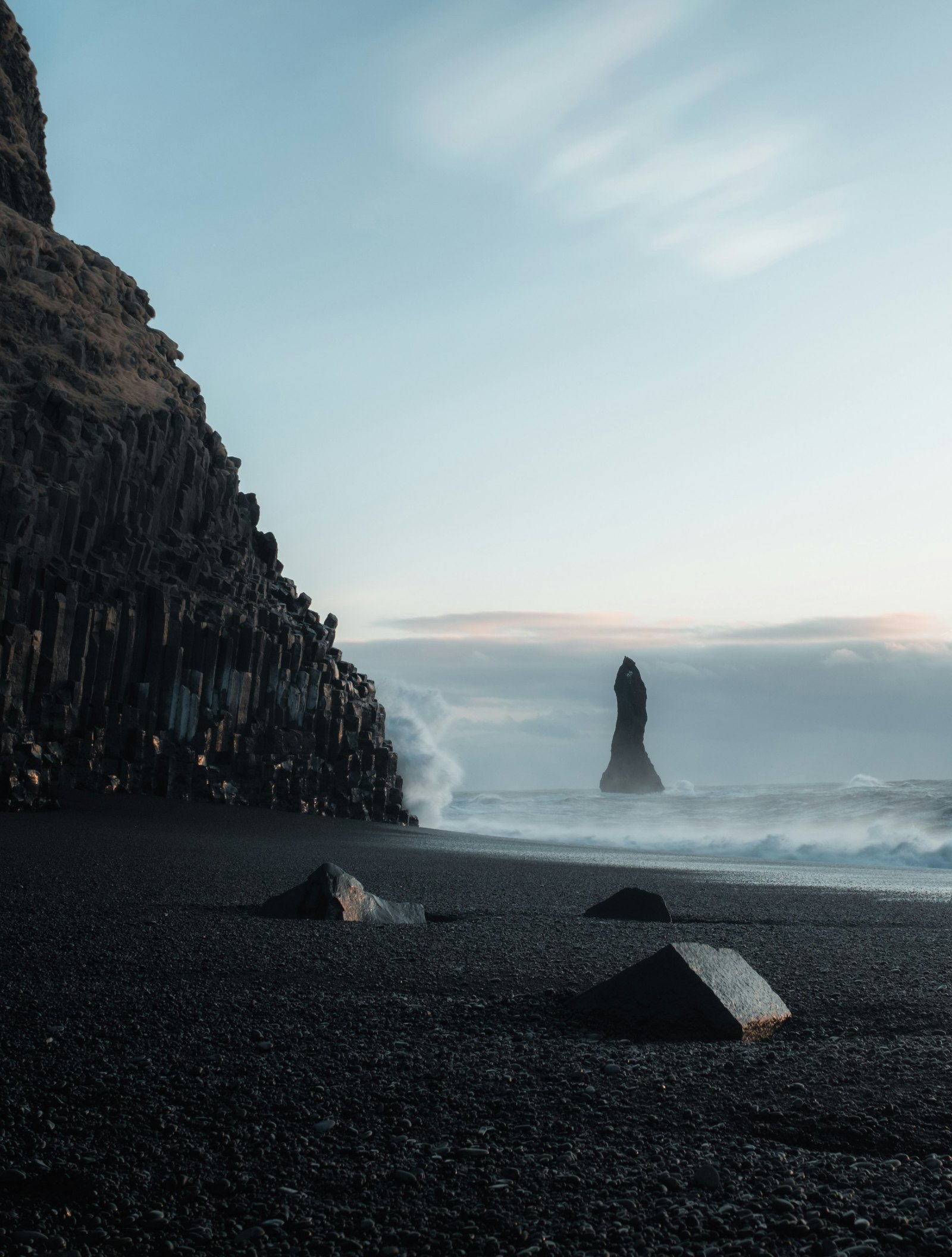 Black sand beach with basalt cliffs and a sea stack offshore, waves breaking under a pale sky.