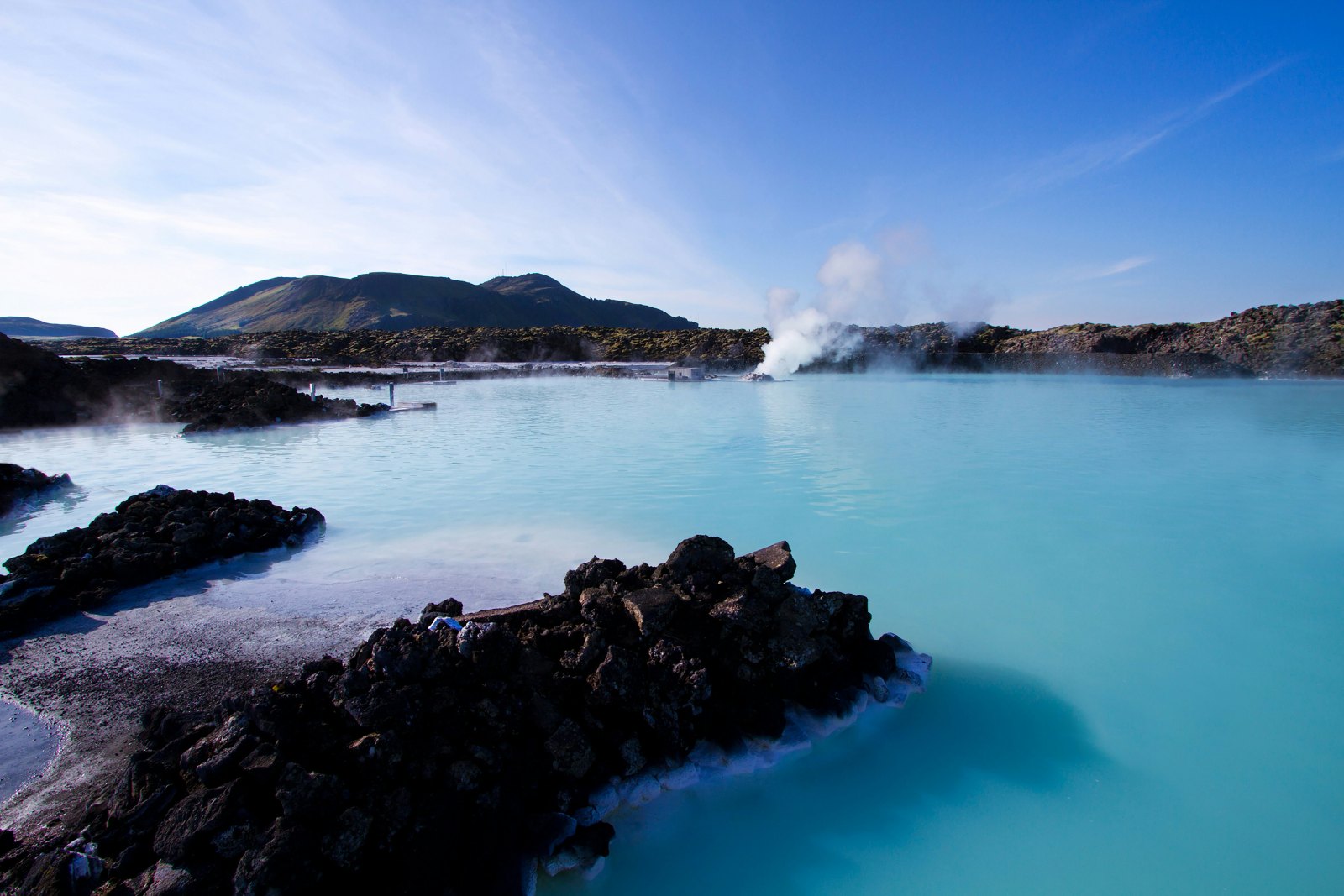 Steam rising from a milky-blue geothermal lagoon among dark lava rocks under a blue sky.