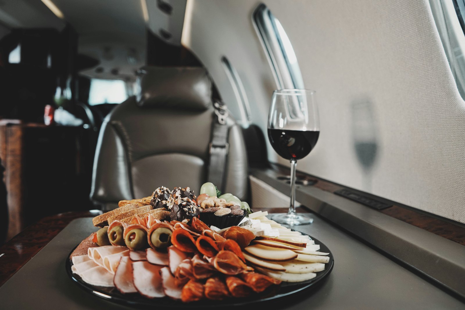 Business jet cabin with a glass of red wine and a platter of food on the table by the window.