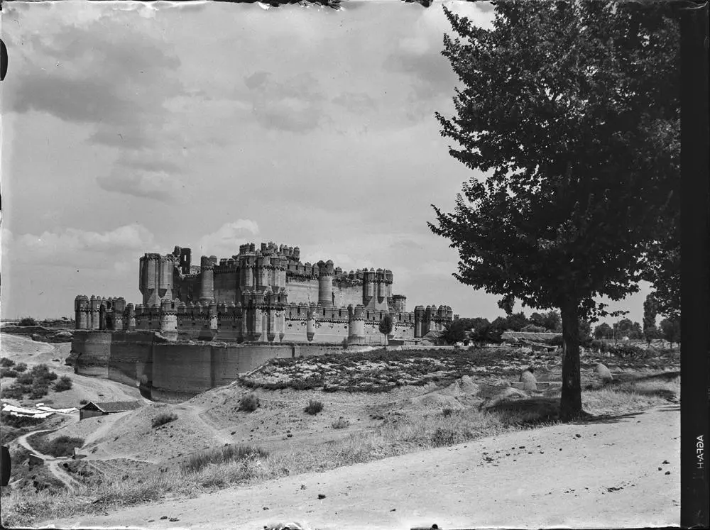 Coca. Castillo [Vista del castillo rodeado por el foso. Destacan las torres que flanquean las cuatro esquinas y su forma almenada. Campos alrededor].