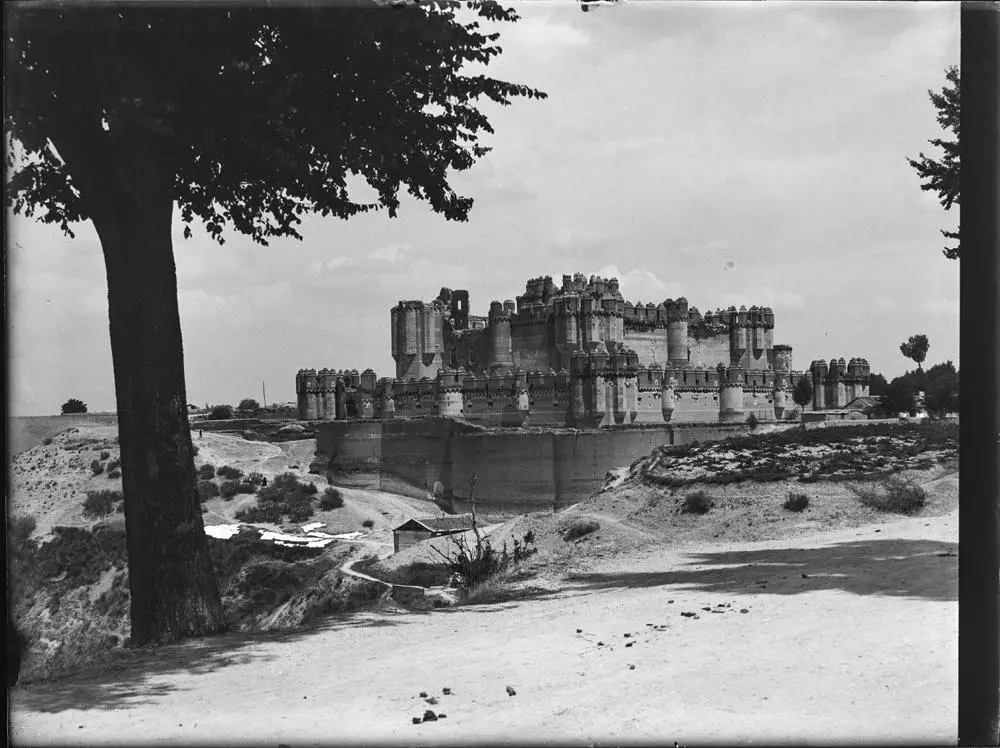 Coca. Castillo [Vista del castillo rodeado por el foso. Destacan las torres que flanquean las cuatro esquinas y su forma almenada. Torre en ruinas. Campos alrededor].