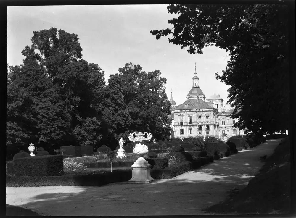 La Granja. Blick auf Schloss v. d. Fuente de la Fama aus [Vista del palacio desde la fuente de la Fama con figuras mitológicas rodeada de árboles y jardines. Ventanas abiertas y cúpula entre los tejados].