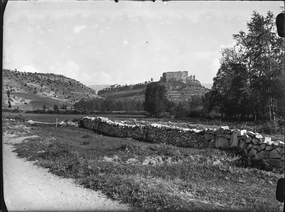 Castillo de Pedraza [Vista del castillo sobre la colina protegido por un muro defensivo. Alrededor, campo y restos de un pequeño muro de piedra bordeando el camino].