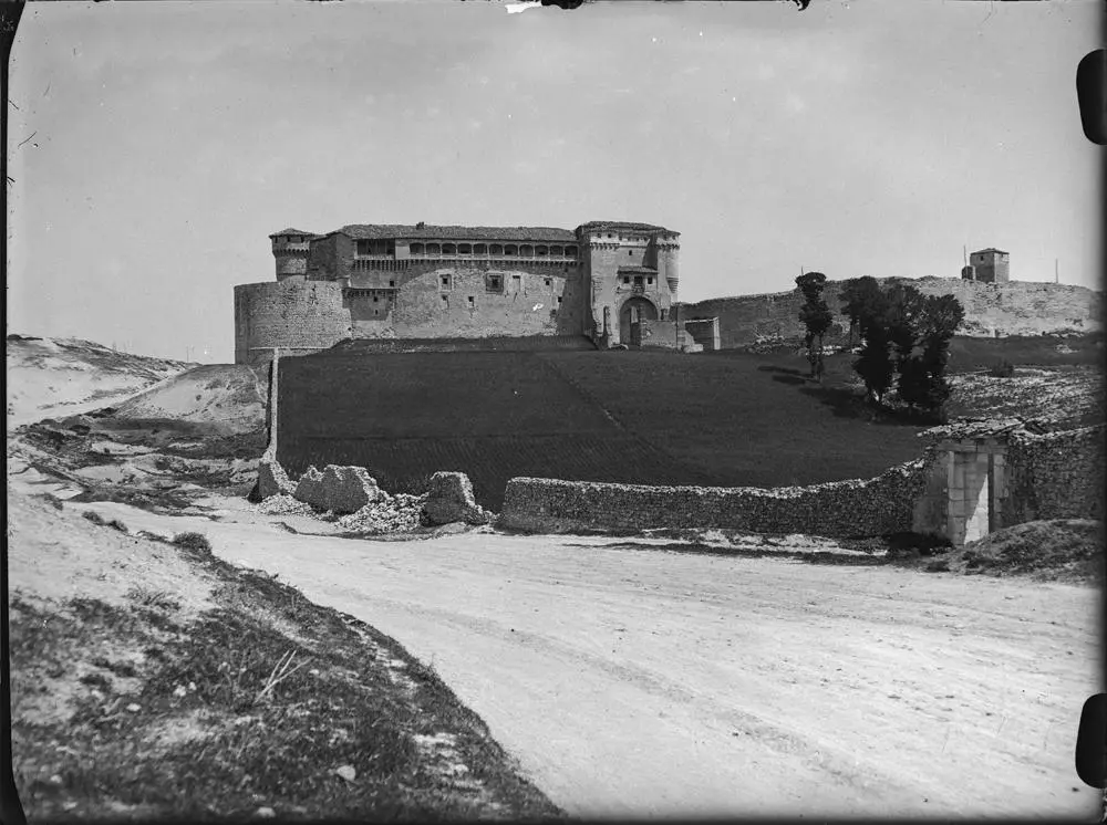 Castillo de Cuéllar [Vista frontal del castillo con campo de cultivo en la explanada delantera rodeado por un muro parcialmente derruido].