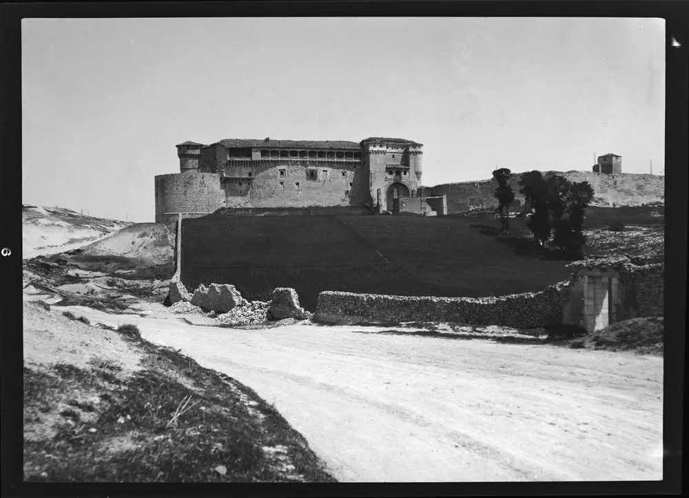 Castillo de Cuéllar [Vista frontal del castillo con campo de cultivo en la explanada delantera rodeado por un muro parcialmente derruido].