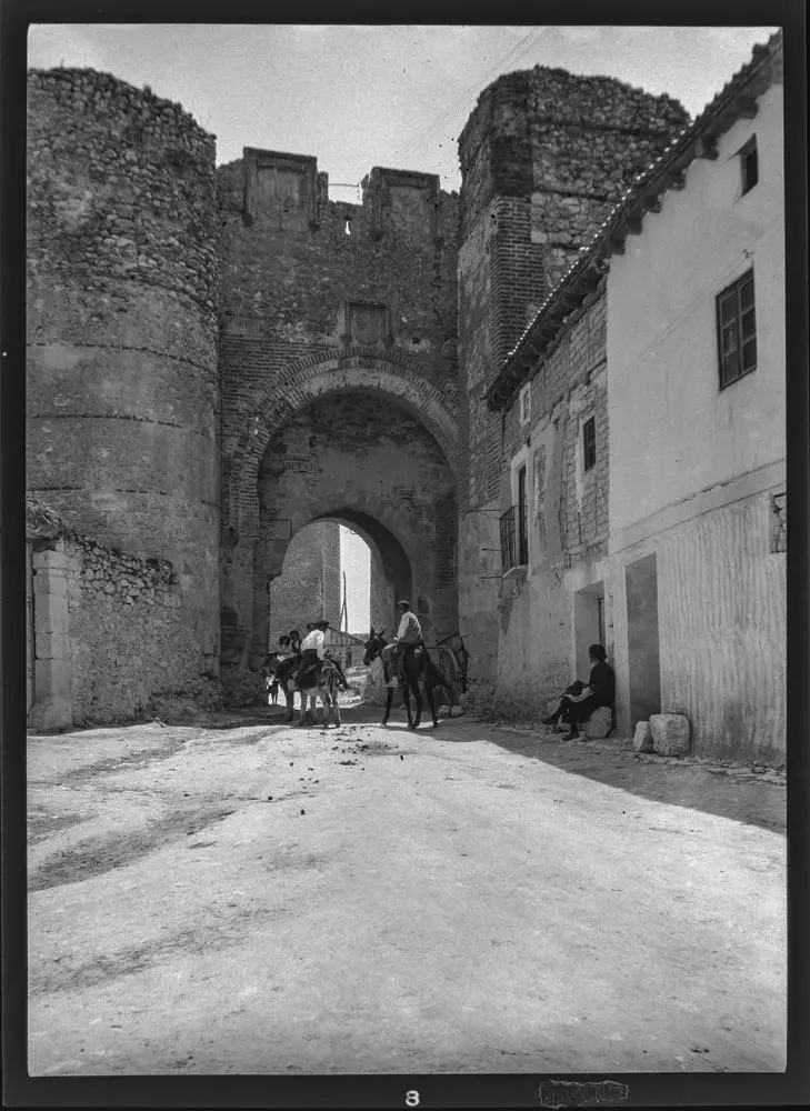 Eingangstor zum Castillo de Cuellar [Puerta de San Andrés flanqueada por una torre cilíndrica y otra rectangular, la acceso al recinto del castillo. Hombres en burro pasando por él y mujer sentada en el banco de una casa].