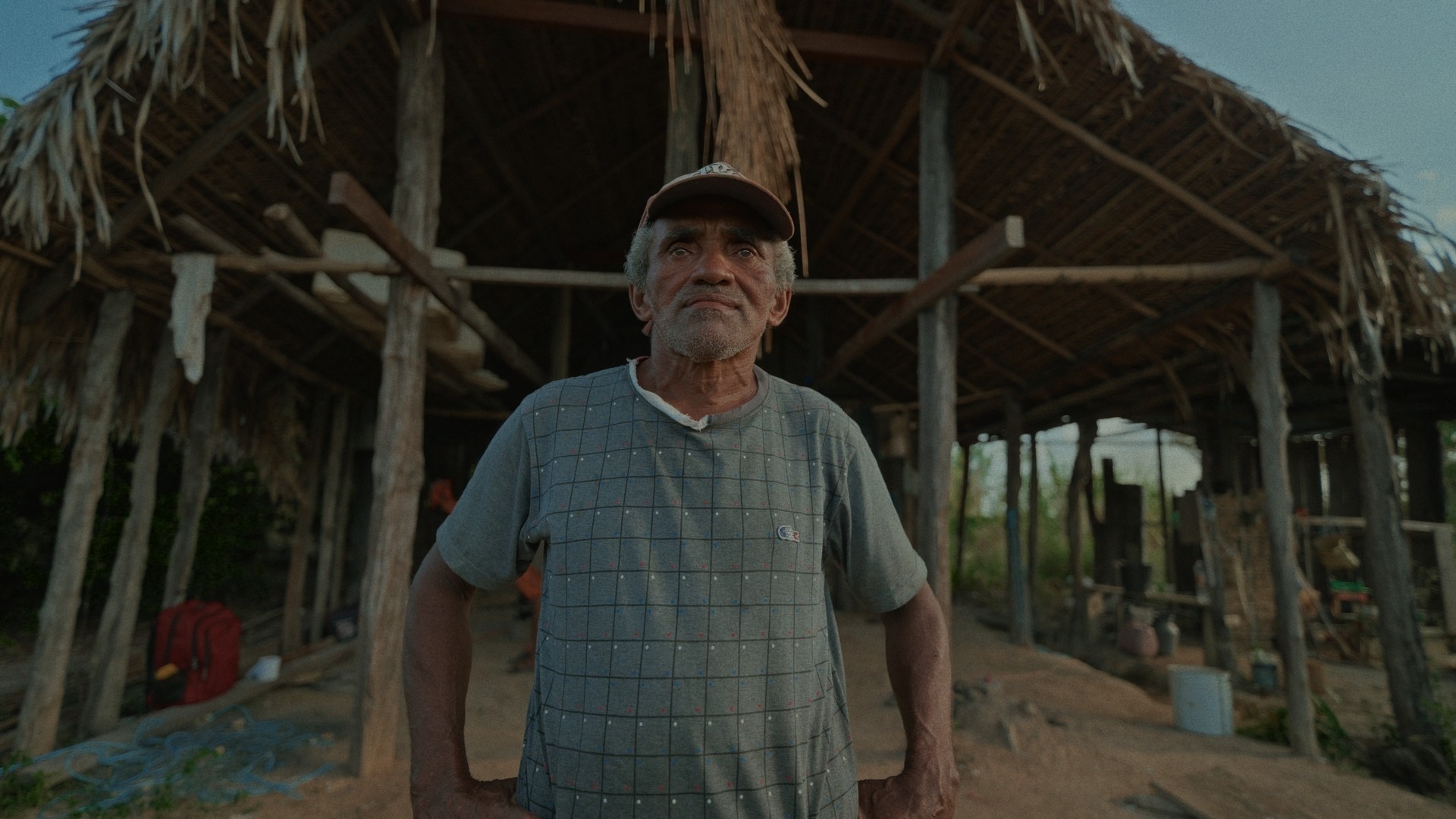 Pescador de meia-idade parado em frente ao seu barracão com telhado de palha e redes para dormir. Vestido com camisa velha cor cinza, calça velha cinza e boné velho cor vermelha com o brasão do Flamengo. É dia e o céu está com poucas nuvens