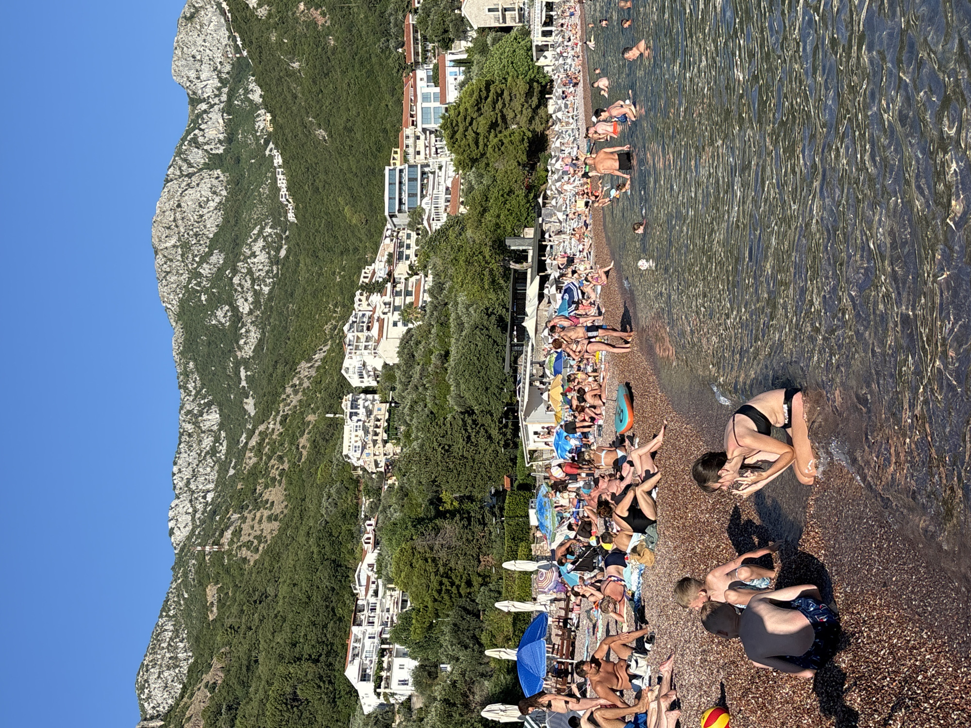 This is a photograph taken at a pebble beach in Sveti Stefan, Montenegro. The scene depicts a crowded beach with numerous people engaged in various summer activities. In the foreground, children and adults are seated on the pebble shore, while others are wading or swimming in the clear, calm water. The sea extends into the background, where more swimmers and boats are visible.
Behind the beach, a dense green landscape rises sharply, with a collection of white buildings, likely hotels and residences, nestled on the hillside. The architecture suggests a resort town. The overall impression is one of a bustling tourist destination during warm weather. The sky is a clear, bright blue, indicating a sunny day. The light suggests it is late afternoon or early evening.