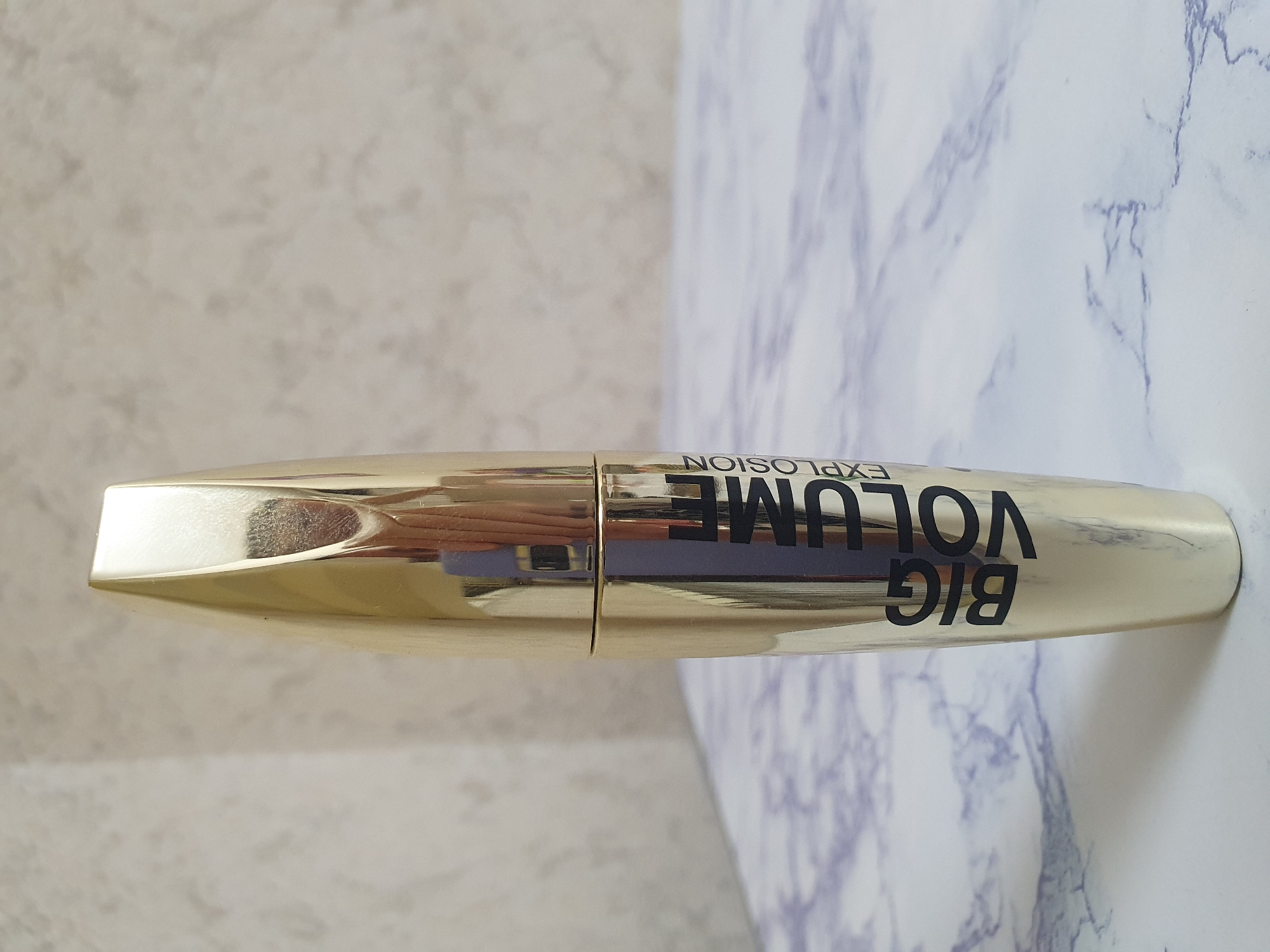 A gold-colored mascara tube stands upright on a marble-patterned surface. The tube has black text that reads "BIG VOLUME EXPLOSION" vertically along its side. The background is a textured, light-colored wall. The image is a still shot, with no people or actions depicted. The environment appears to be an indoor setting. The location in Tashkent, Uzbekistan, is not discernible from the image itself.