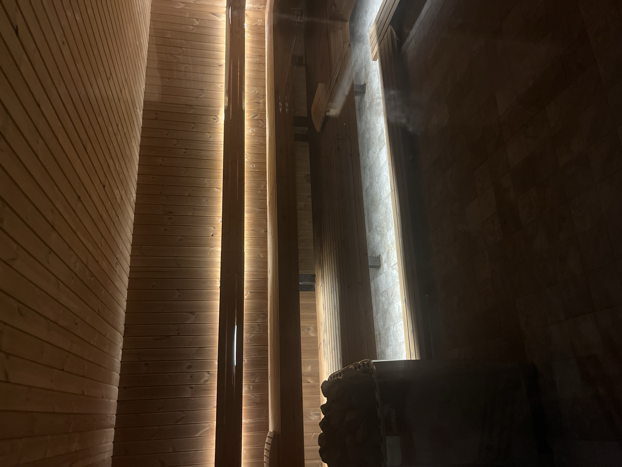 The image depicts the interior of a sauna. Walls are constructed of light-colored horizontal wooden planks. Vertical wooden beams are positioned to create illuminated recessed channels. A dark stove sits in the foreground. The location is not discernible from the image and information about the city of Sofia, Bulgaria is not identifiable.