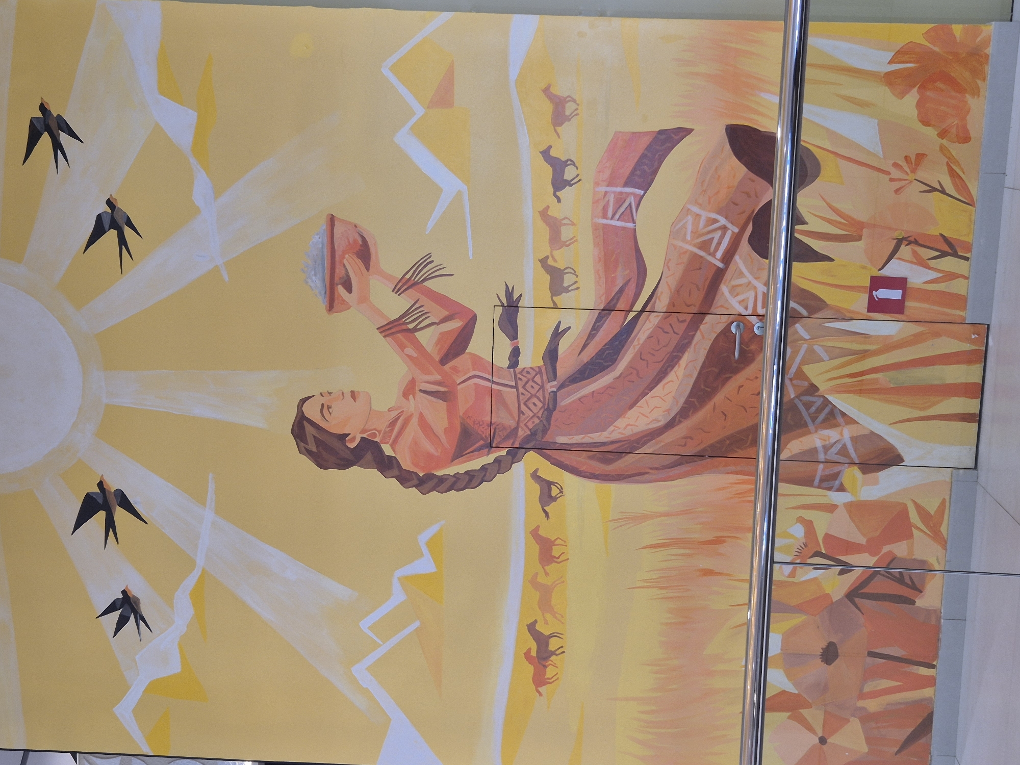 The image shows a mural on a wall in Kokshetau, Kazakhstan. The mural features a woman in traditional Kazakh clothing, holding a bowl. The background is a yellow and orange landscape with geometric mountains, birds, horses, and a stylized sun. A metal pole partially obstructs the right side of the mural, and there are rectangular panels below the main image with geometric floral patterns. A sign indicating the location of the water closet (WC) is also visible.