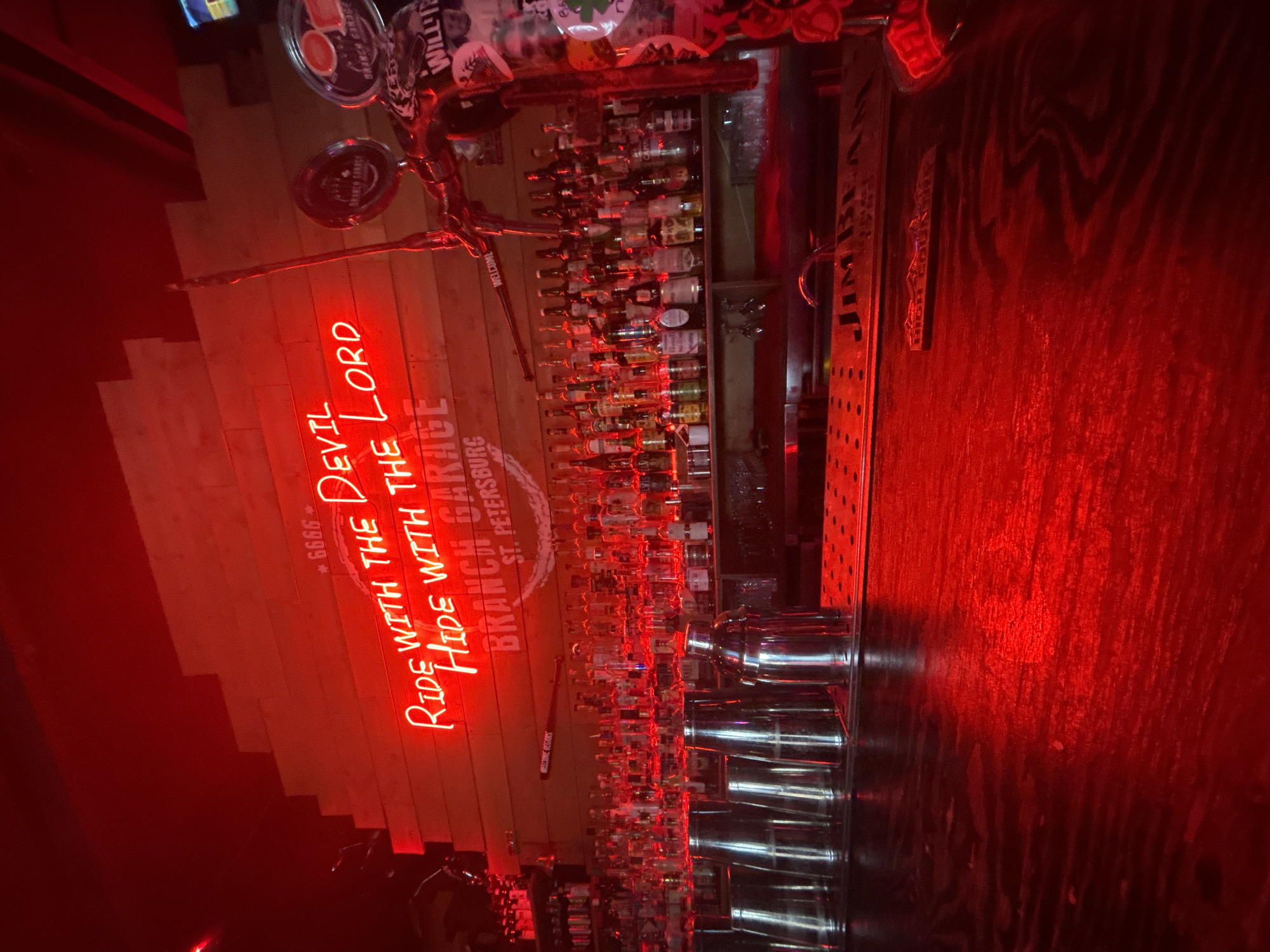 The image shows the interior of a bar in St. Petersburg, Florida, specifically the "Branch Garage" location. The bar has a rustic, perhaps biker-themed aesthetic, evidenced by the neon sign that reads "Ride with the Devil Hide with the Lord" and the phrase "Branch Garage St. Petersburg" underneath. There's a wall covered with bottles of various liquors and spirits, a bar counter with cocktail shakers lined up. The environment is dimly lit with a strong red hue, which may be from the neon sign.
