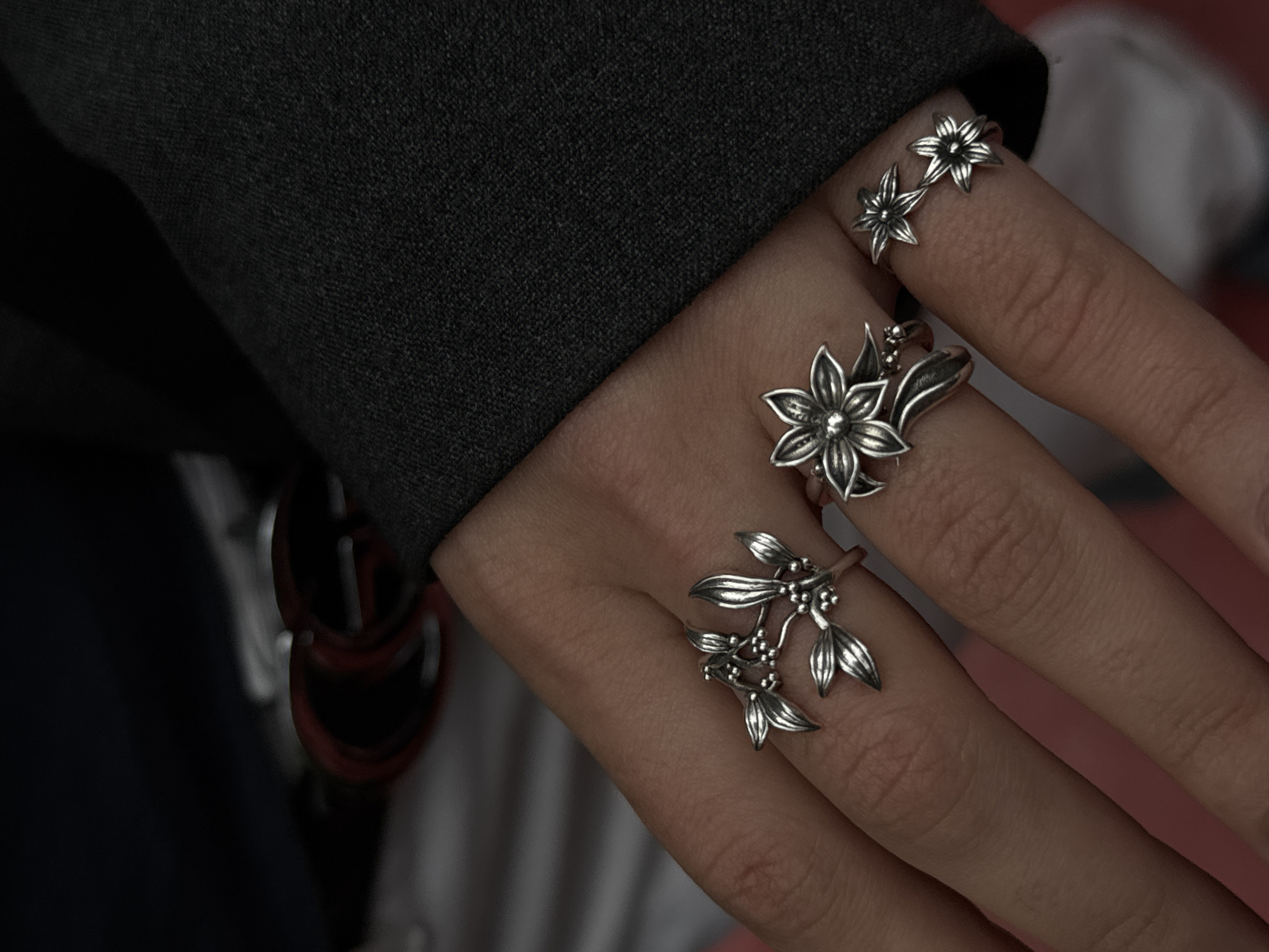 The image shows a close-up of a hand adorned with three silver rings, each featuring a floral design. The rings are positioned on the pinky, ring, and middle fingers. The hand, with fair skin, is placed against a dark gray textured fabric, likely clothing. In the background, blurry white and red colors suggest additional clothing or items. The specific location in Yerevan, Armenia, is not discernible from the provided image.