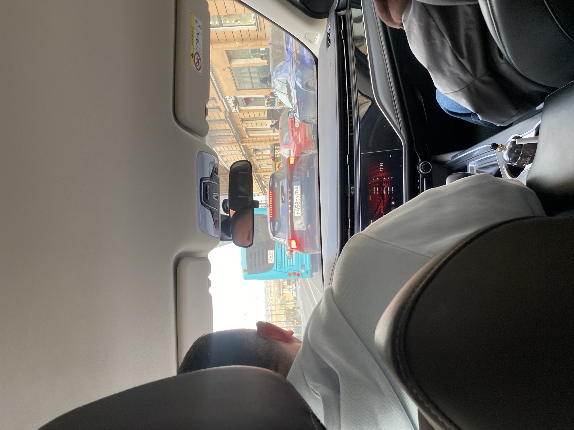 The photograph was taken from the interior of a car in Saint Petersburg, Russia. The car is stopped in traffic. Other cars and a blue bus are visible through the windshield. The license plate of the car directly in front reads "У 458 СУ 198." A person is seated in the back seat, wearing a white hoodie. The head of the person is visible over the back of the car seat. The front dashboard and central console are visible. The time is 17:19, according to the digital display.
