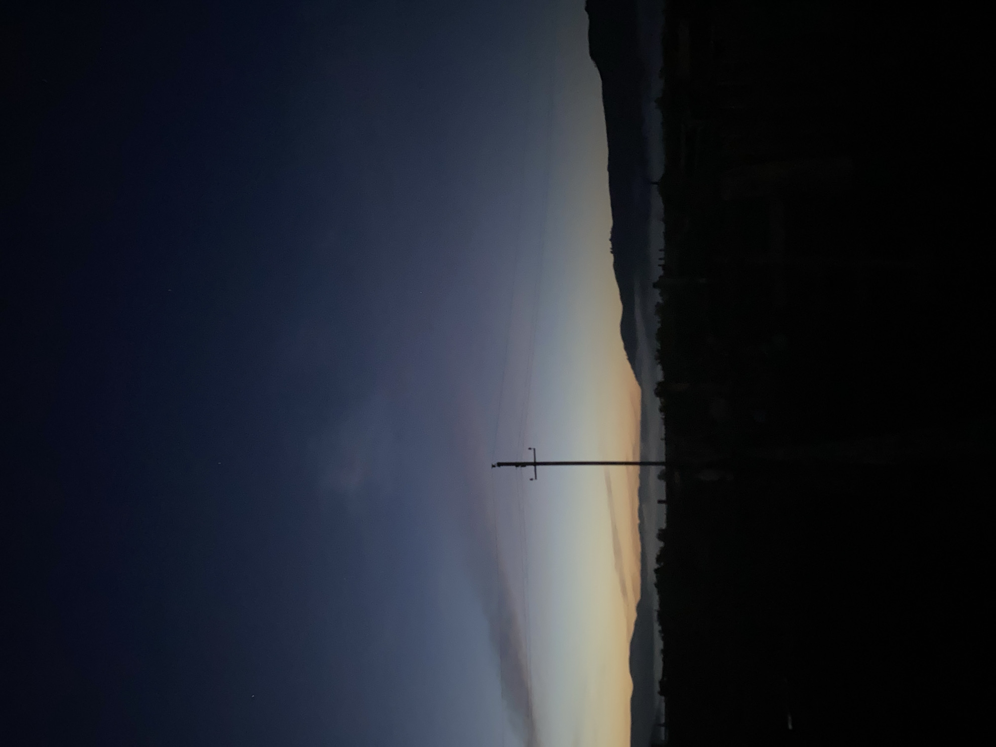 The image captures a pre-dawn or post-dusk scene in Maloarslangulovo, Russia. The sky is a deep, dark blue that transitions to lighter shades of blue and a hint of orange near the horizon, suggesting the presence of either sunrise or sunset. Faint clouds are visible in the upper portion of the sky. A lone utility pole stands prominently in the middle ground, with wires stretching horizontally across the frame. In the distance, a silhouetted landscape of rolling hills or mountains is visible, partially obscured by a low-lying mist or fog that hugs the terrain. The foreground is entirely black, indicating it is in shadow. No people, animals, or specific structures are clearly discernible due to the low light conditions. The overall atmosphere is one of quiet solitude and natural beauty during twilight.