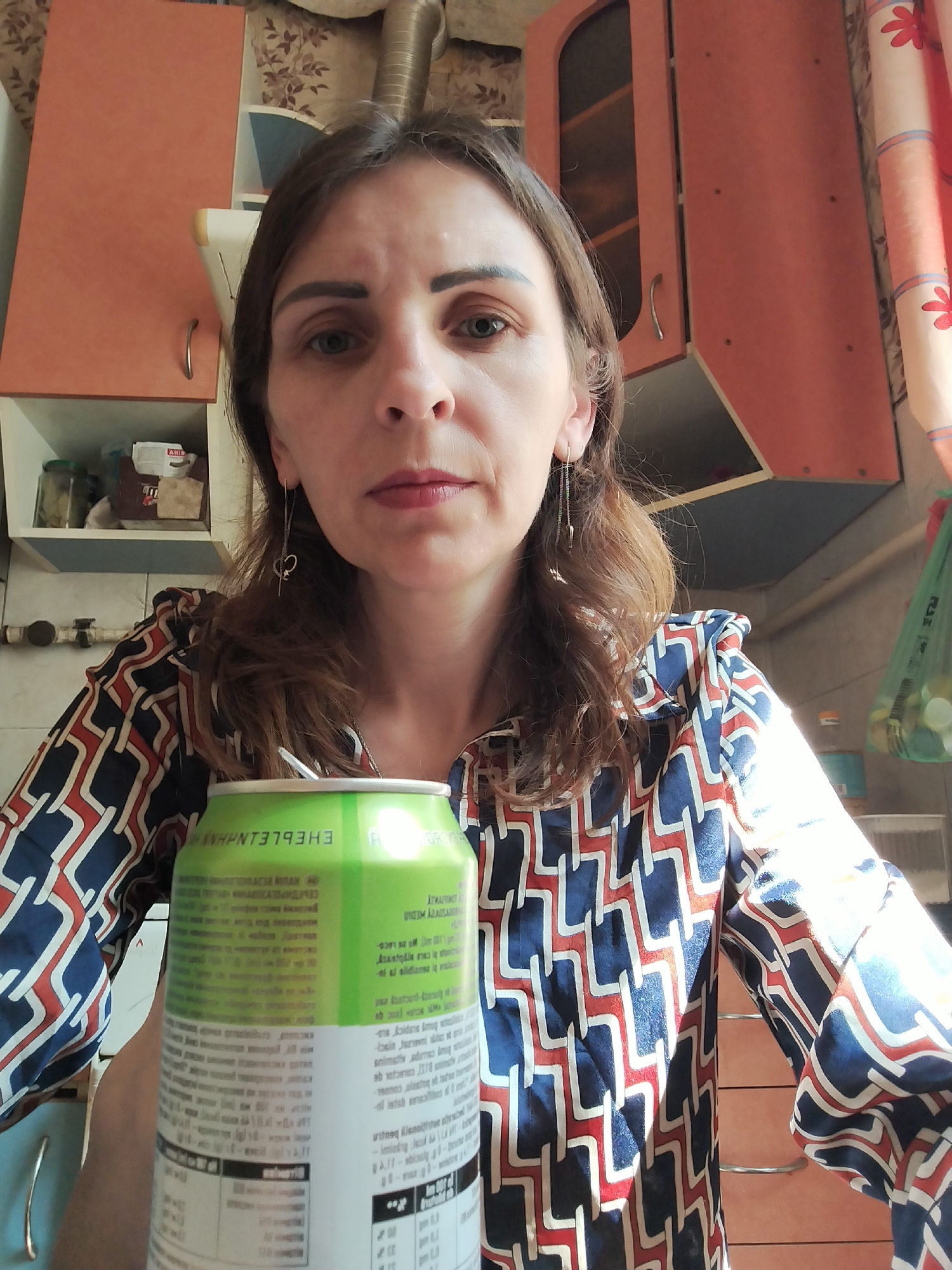 A medium shot shows a woman with shoulder-length brown hair standing in what appears to be a kitchen. She is looking directly at the camera with a neutral expression. She is wearing a collared shirt with a geometric pattern of navy blue, white, and reddish-orange. Her left arm is holding up a green can of energy drink, with text on the can partially visible. The background shows kitchen cabinets, a shelf with jars and containers, and some curtains. The overall setting suggests an indoor domestic environment. There are no clear indicators of the city or country in the image.