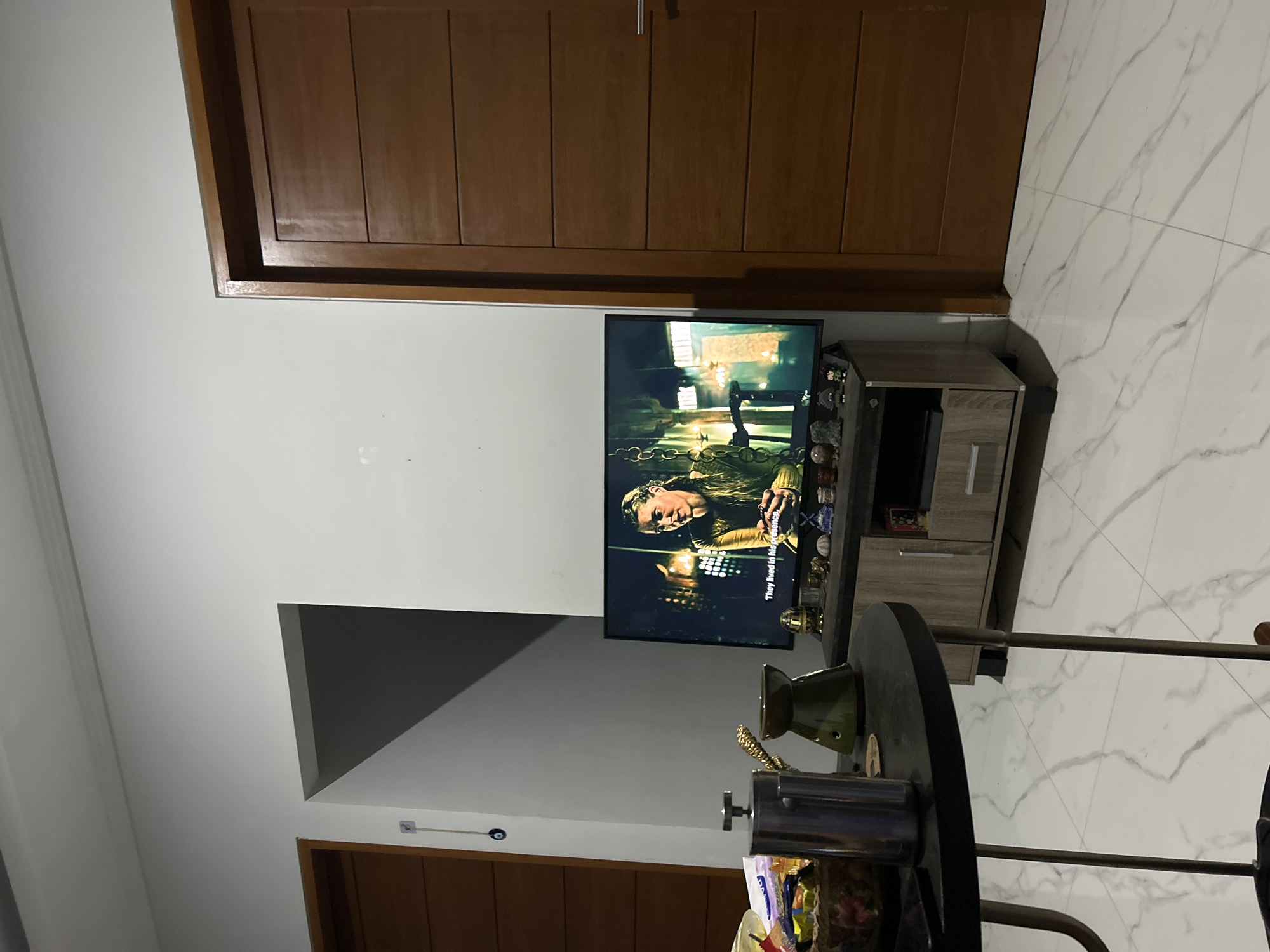 The image captures a home entertainment setup in what appears to be a living room. A flat-screen television is mounted on a wall, displaying a scene with a person dressed in historical attire. The TV is placed above a small, light-wood entertainment cabinet. On top of the cabinet, a collection of decorative items, possibly figurines or small sculptures, are arranged. To the right of the television, white tiled flooring with subtle grey veining is visible.
In the foreground, a dark, round table is partially visible, with a metallic coffee pot or dispenser and some packaged snacks on it. The room's walls are painted white, and a large wooden door with vertical paneling is seen on the left side of the image. Another similar wooden door is partially visible at the bottom. The overall lighting suggests it is daytime. The scene appears calm and domestic, typical of a residential space. There is some text on the TV screen, "They lived in his presence." The location context provided is Sukawati, Indonesia.