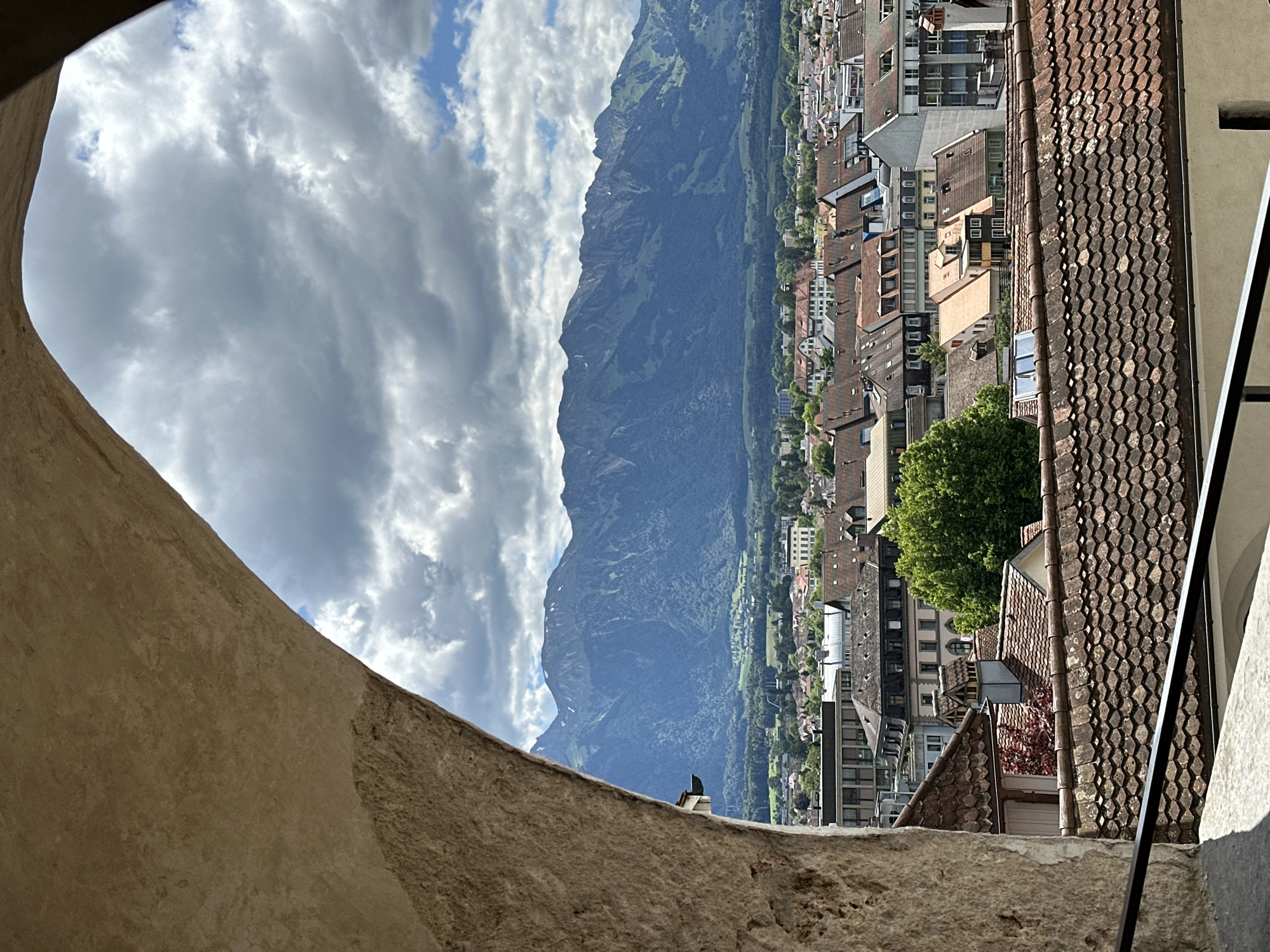 The photograph captures a scenic view of Thun, Switzerland, framed by a rustic stone archway in the foreground. The arch, made of rough-hewn stone, dominates the left and upper portions of the image, with its textured surface suggesting age and solidity. Through the arch, a panoramic vista unfolds.
The middle ground is occupied by a dense cityscape of Thun, characterized by a sea of terracotta-tiled roofs, indicative of traditional European architecture. Various buildings, ranging from multi-story structures with classical facades to more modern designs, are visible. A prominent, vibrant green tree stands out amidst the urban sprawl, providing a splash of natural color.
Beyond the city, a majestic mountain range forms the backdrop. These mountains are steep and covered in lush green vegetation on their lower slopes, transitioning to rocky, snow-dusted peaks at higher elevations. The sky above is a dynamic mix of dramatic, dark grey clouds interspersed with patches of bright blue sky, suggesting an overcast but potentially clearing day. The overall impression is one of a historic Swiss town nestled against a dramatic natural landscape.