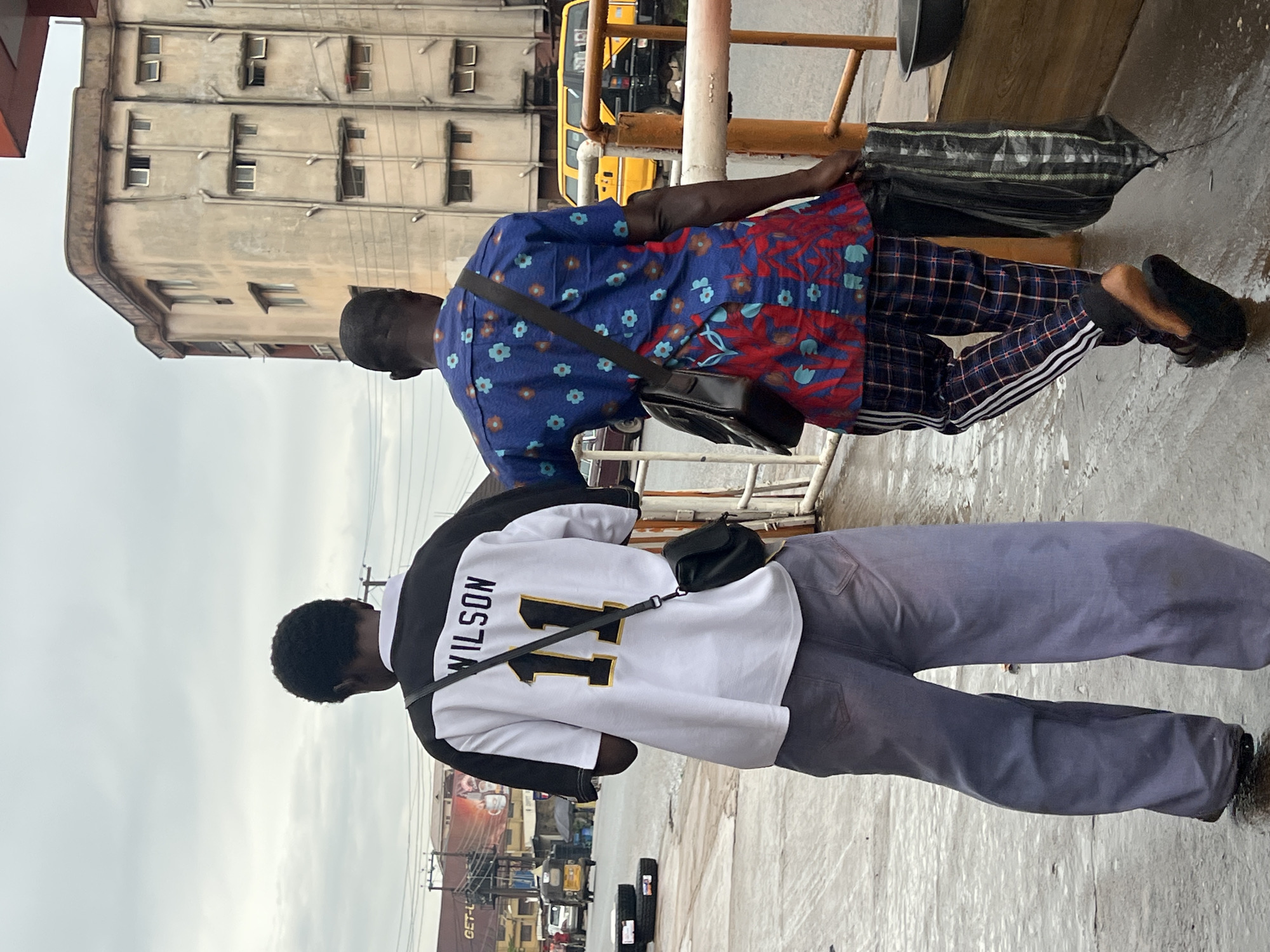 The image shows two men standing on a wet, possibly concrete, surface. The man in the foreground, closer to the camera, is wearing a white and black long-sleeved shirt with "WILSON" and the number "11" printed on the back in black and gold. He has a black crossbody bag. He is wearing grey trousers. The second man is further away, wearing a blue and red patterned short-sleeved shirt and checkered trousers with white stripes down the side. He also has a black crossbody bag. Behind the men, there is a building with multiple windows, a yellow bus, and some metal railings. The scene appears to be outdoors, possibly in an urban or semi-urban area, given the building and the bus. The ground is wet, suggesting it might have recently rained or there's a water source nearby. The sky is overcast, indicating a cloudy day. There is text visible on the shirt of the man in the foreground: "WILSON 11". There are also some signs of shops and advertising in the background, some with text that is not clearly legible. The overall mood seems neutral, with the men appearing to be in transit or waiting. The context provided places this scene in Nkpor, Nigeria.