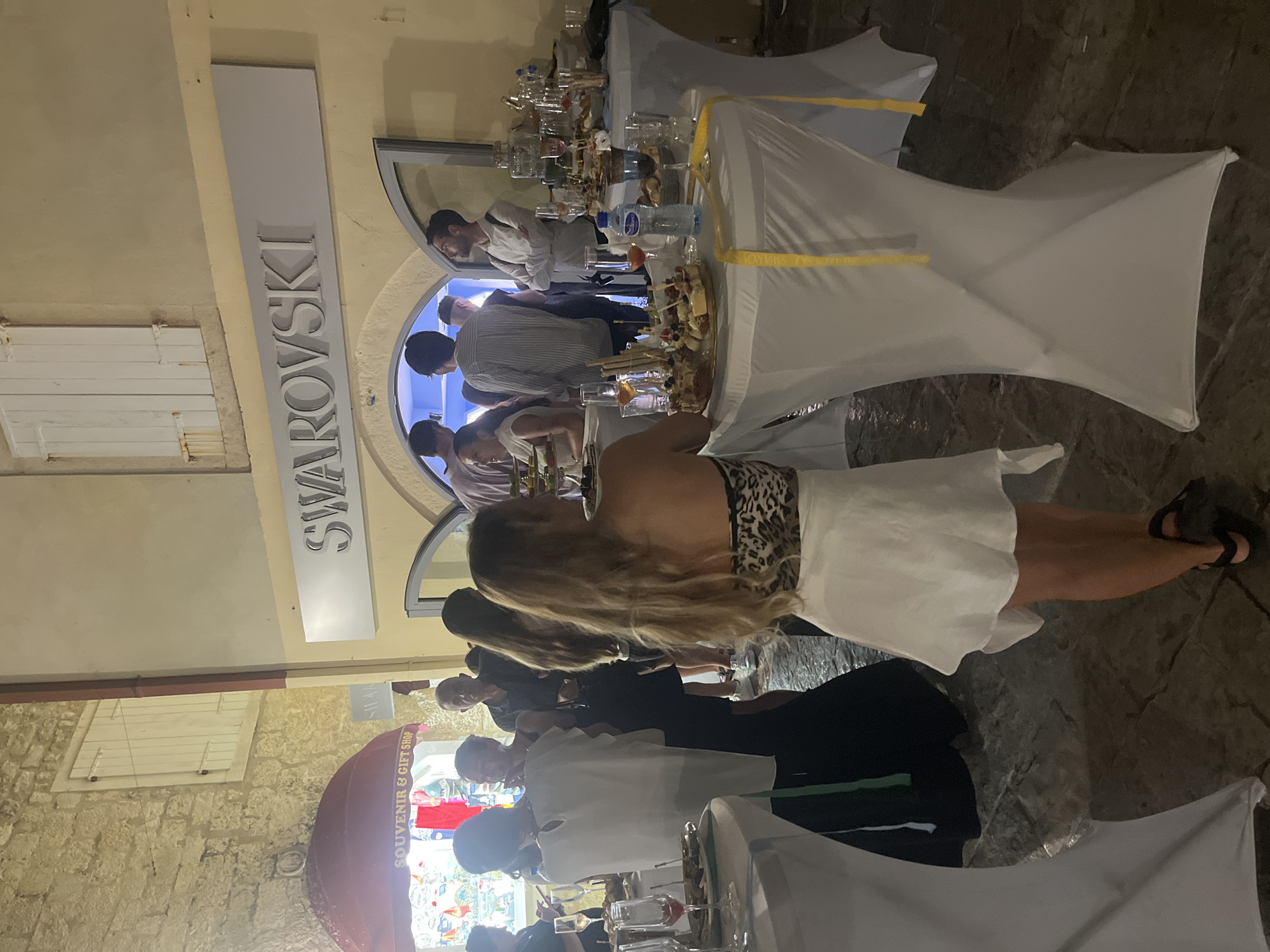 The image depicts a social gathering outside a Swarovski store in Budva, Montenegro. Several individuals are observed mingling, with some standing near tables laden with food and drinks, while others are engaged in conversation or observing the surroundings. A sign above the store entrance clearly reads "SWAROVSKI". White tablecloth-covered tables are arranged, displaying platters of appetizers, glasses, and bottles of beverages, including a prominent bottle of water. The ground appears to be paved with cobblestones, suggesting an outdoor urban setting. The overall atmosphere suggests an event or reception held in the evening.