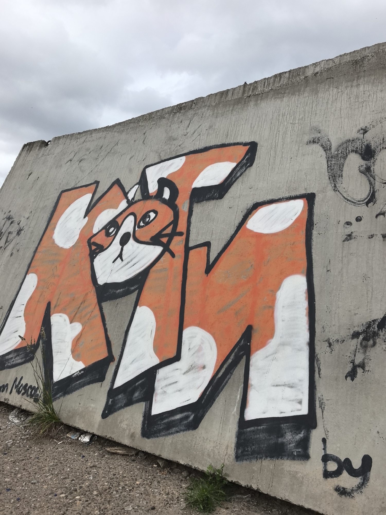 The image shows graffiti on a concrete barrier, likely in the city of Chaikovsky, Russia. The graffiti features large, orange and white letters forming the word "ATM," with a stylized cartoon fox face integrated into the "A." The fox has wide eyes, a black nose, and whiskers. The concrete barrier itself appears weathered, with streaks of discoloration and other, less prominent black graffiti art on its surface. The ground in front of the barrier is a mixture of gravel and dirt, with some sparse vegetation growing along the base. The sky above is overcast with gray clouds. The word "by" is scrawled in black graffiti to the right of the main artwork, suggesting a tag or artist's signature.