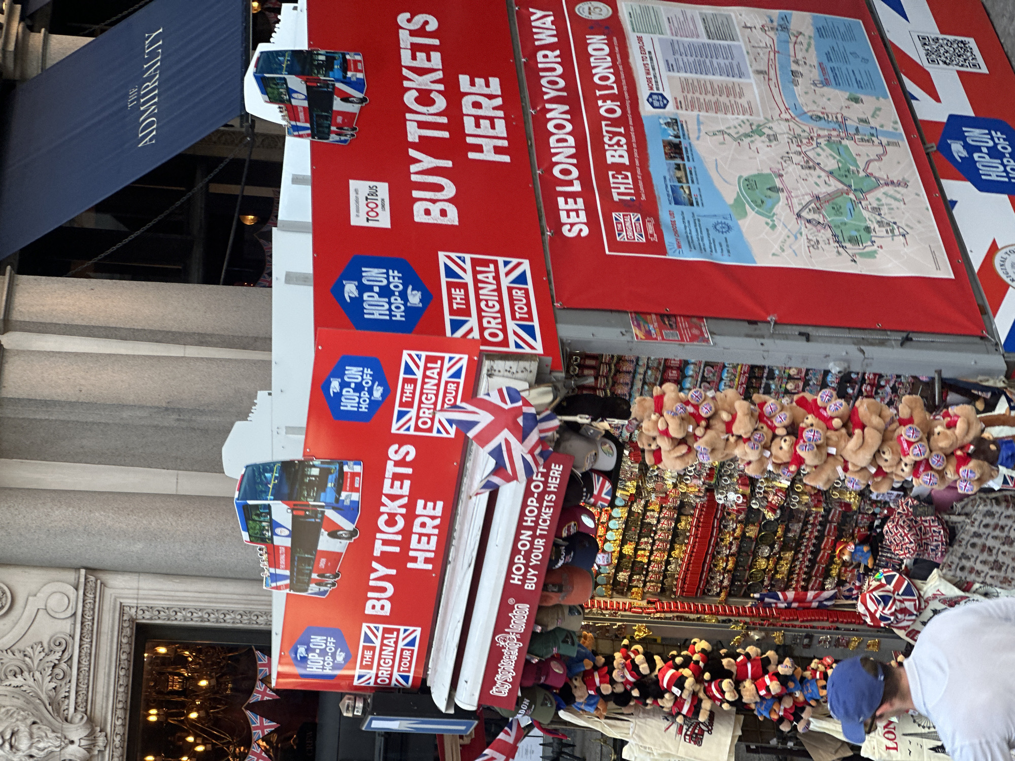 The image depicts a street scene in London, United Kingdom. A red banner with "BUY TICKETS HERE" and "SEE LONDON YOUR WAY" is prominently displayed, advertising "The Original Tour" hop-on-hop-off bus service. Souvenir items such as teddy bears, hats, and magnets adorned with British flags are on display for sale. The "Admiralty" sign can be seen in the upper left corner of the image, indicating the location is near Admiralty Arch. People are present in the scene, including a person wearing a blue baseball cap. The environment suggests a bustling tourist area.