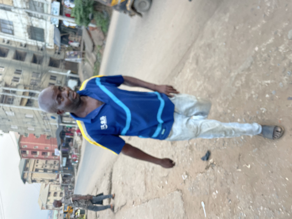 An outdoor, daytime photograph captures a man standing on an unpaved, dusty path next to a road, with buildings and another person in the background. The image is taken from a slightly low angle and is rotated counter-clockwise.
The primary subject is a bald, dark-skinned man of apparently middle age or older, looking towards the viewer's left side. He wears a blue short-sleeved polo shirt with distinctive light blue and yellow stripe designs across the chest and shoulders. On the left side of his chest, a white logo or text that appears to be "3B" is visible. He is also wearing light-colored pants, which appear somewhat soiled or worn, and dark sandals or flip-flops on his feet. His expression is neutral or slightly serious. His left arm is bent at the elbow, while his right arm is mostly obscured but appears to be by his side.
The ground where he stands is a mixture of dirt and gravel, with visible debris including a white plastic spoon and a dark, unidentified object that might be a discarded slipper or piece of trash. To his right and behind him, a section of a paved or hard-packed dirt road is visible. Further down this road, another person can be seen walking, smaller in the frame, wearing a patterned shirt and dark pants.
In the background, on the left side of the frame, a cluster of multi-story buildings stands, suggesting an urban or semi-urban environment. Some buildings are light-colored (grey or off-white), while one in the mid-ground is a reddish-brown hue. Visible electrical wires or pipes run along the exterior of some structures. Small commercial establishments or signs are faintly discernible near the buildings. A part of a yellow vehicle, possibly a tricycle or rickshaw, is visible at the far upper right edge of the frame. The sky is light, indicating daylight conditions.