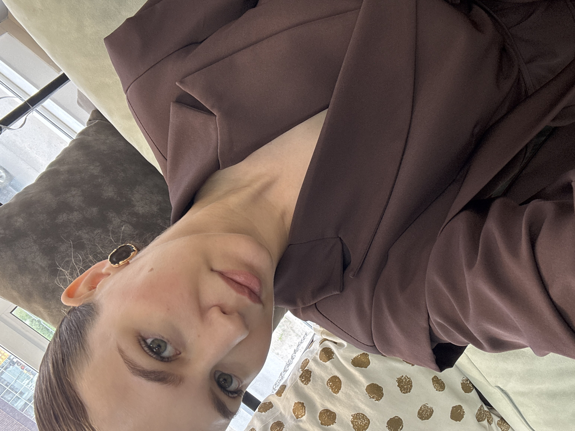 A woman is taking a selfie in a room with natural light. She has her hair pulled back and is wearing a dark brown blazer. She has on large, oval-shaped earrings with a dark stone and a gold rim. In the background, there are several pillows and a window with blinds. The room appears to be a living space, possibly an apartment. The presence of a window suggesting an urban setting, and the type of interior decor, is consistent with an apartment in Chaikovsky, Russia.