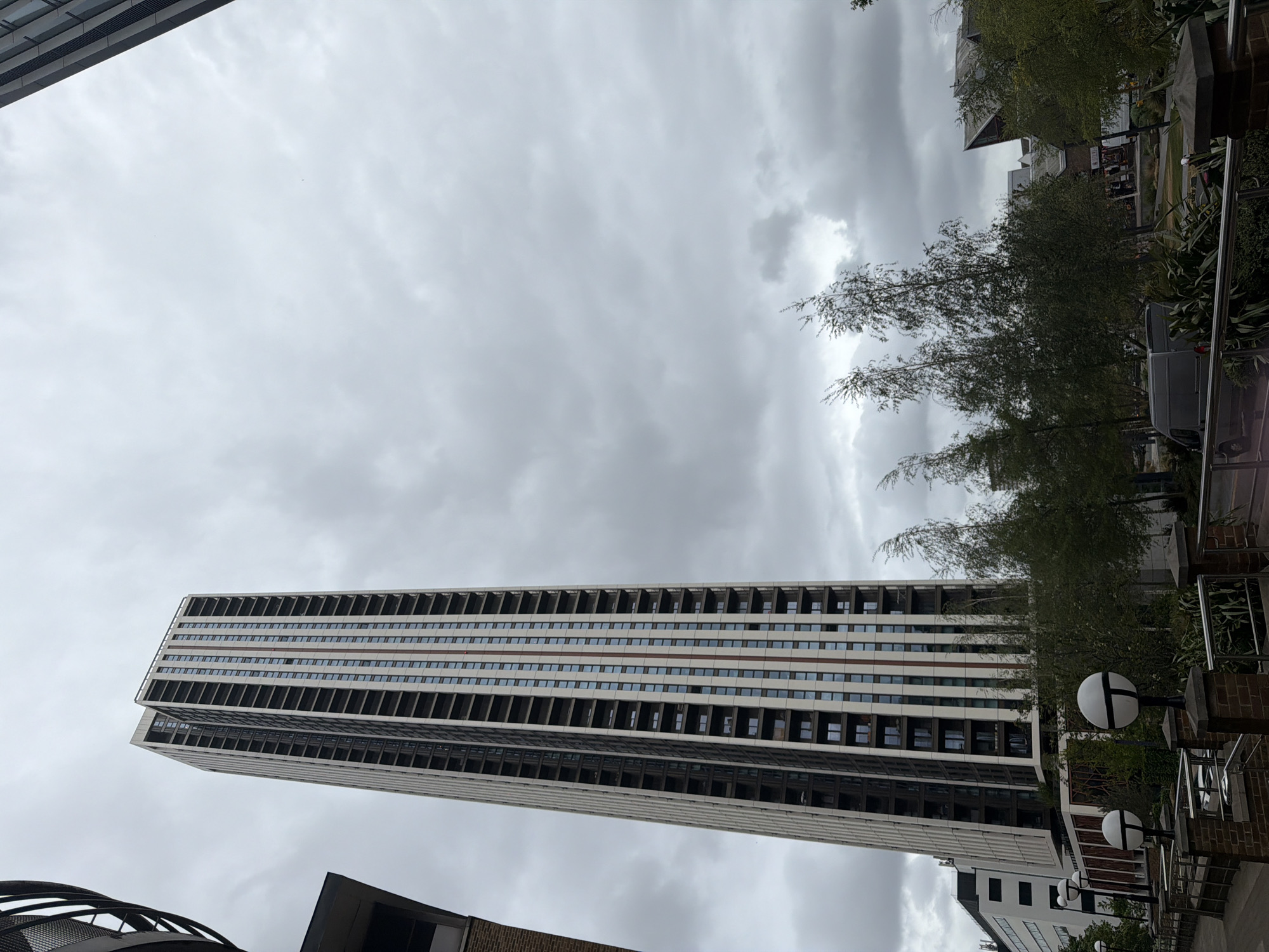 A tall, rectangular building dominates the frame against a cloudy sky. The building has a repetitive pattern of horizontal lines and window openings. Adjacent to the building are trees and greenery, along with street lamps and elements of urban infrastructure. The scene is set in an urban environment, possibly the city of London, United Kingdom. The details of the architecture and surrounding environment suggest a modern cityscape.
