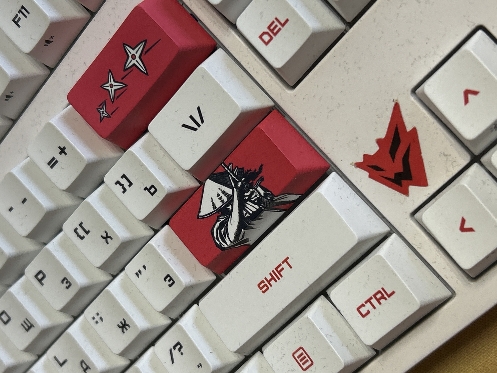 The image shows a close-up of a white and red custom mechanical keyboard. Keycaps have red legends. Some are red and decorated with symbols or stylized images. Keycaps visible include "DEL", "SHIFT", and "CTRL", plus a "Star" and a "Samurai" design. Part of a keyboard case is visible with red decorative design. Other keys are also visible with Cyrillic letters and symbols. The image provides no information to establish the location of the keyboard.