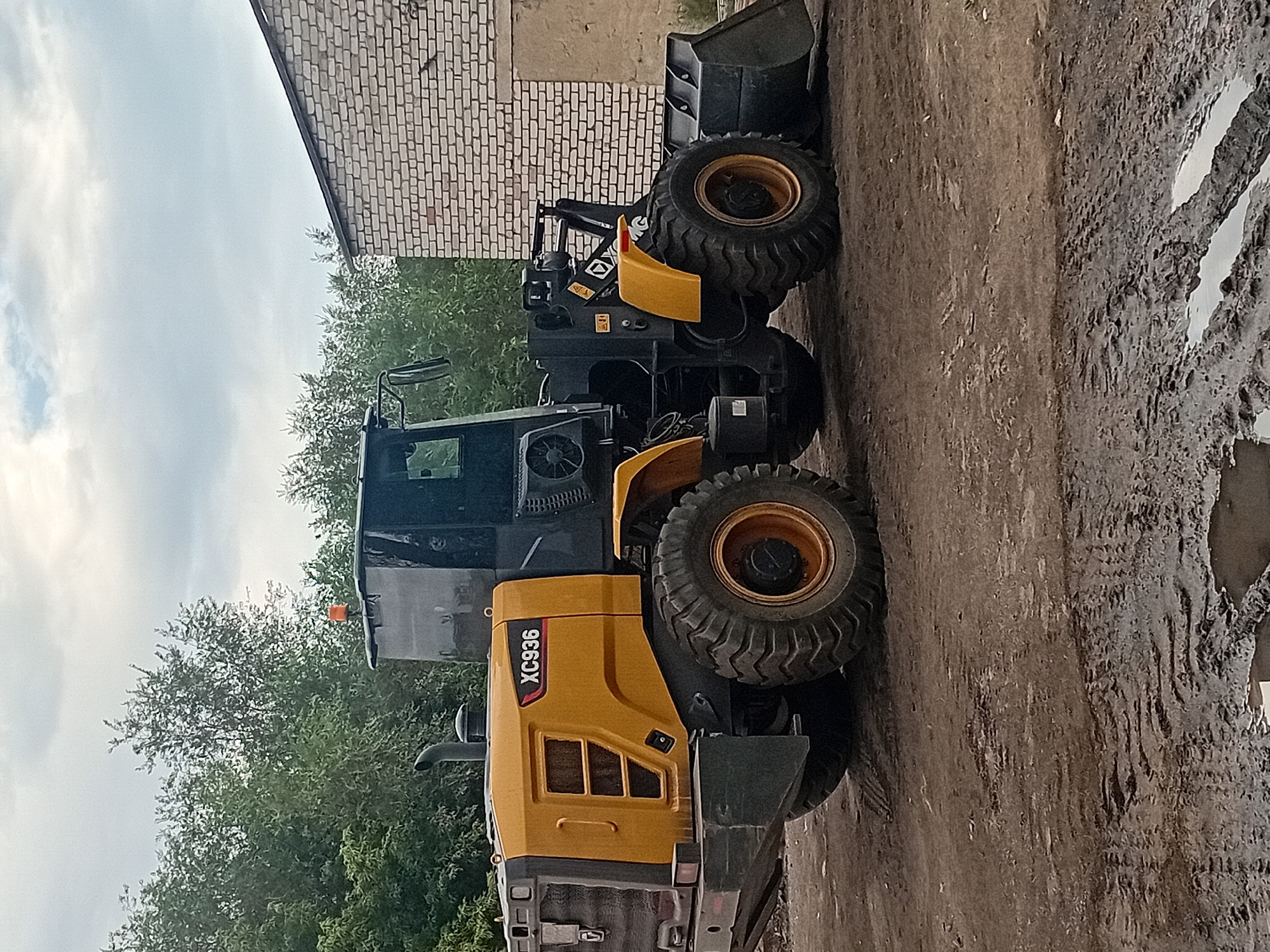 This is a full, eye-level, slightly angled shot of a yellow and black wheel loader parked on a muddy dirt surface. The loader is the primary subject, occupying the majority of the frame. It has a large bucket at the front, heavy-duty tires, and a cab with tinted windows. The model number "XC936" is visible on the side of the loader. In the background, there are green trees and a partially visible brick building. The sky is overcast with clouds. The scene appears to be an outdoor work area or storage yard, possibly in an industrial or construction setting. There are no people or animals present. The overall impression is one of industrial equipment in an outdoor environment. The location in Semey, Kazakhstan is not explicitly identifiable from the visual information alone; this detail would require external context or knowledge of the specific area depicted.