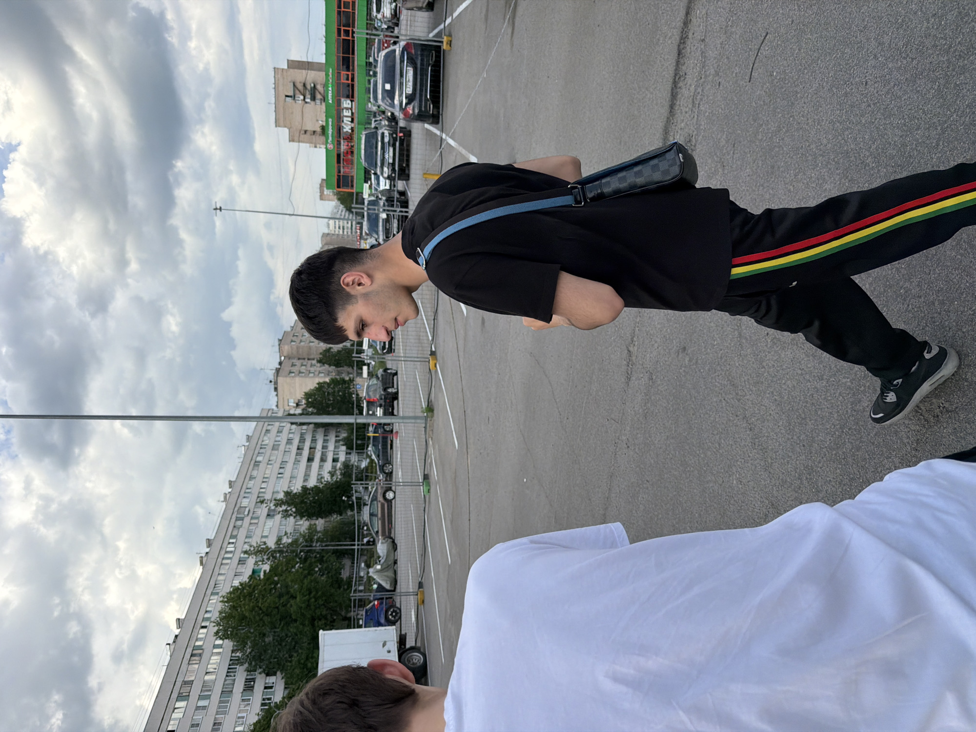 The image displays an outdoor urban scene, likely a street or parking lot in Saint Petersburg, Russia, as suggested by the architecture and visible Cyrillic text. The sky is overcast with large, fluffy clouds, indicating daytime.
In the mid-ground, a young man is visible from his right side, appearing to be walking or standing. He has dark, short hair and is wearing a plain black t-shirt, black track pants with distinct red, yellow, and green stripes running down the sides, and black sneakers with white soles. A dark, checkered shoulder bag with a blue strap crosses his body. His head is tilted downwards, and his expression is neutral.
Partially visible in the foreground, on the right, is the back of another person's head and shoulder, dressed in a white t-shirt.
The background features several buildings and parked vehicles. To the left, a large, multi-story apartment building, consistent with Soviet-era architectural styles, extends across the horizon. To the right, behind a row of cars, stands a commercial building with a prominent green facade. This building displays Cyrillic text, including "АРТЕЛЪ" (Artel), "ХЛЕБ" (Khleb, meaning bread), "Почта России" (Pochta Rossii - Russian Post), and "СДЭК" (SDEK - a delivery service), confirming the Russian context. Numerous cars are parked in what appears to be an asphalt parking lot, marked by white lines and separated by yellow posts and fences. Streetlights or poles are scattered throughout the scene. The overall atmosphere is that of a typical urban setting under a cloudy sky.