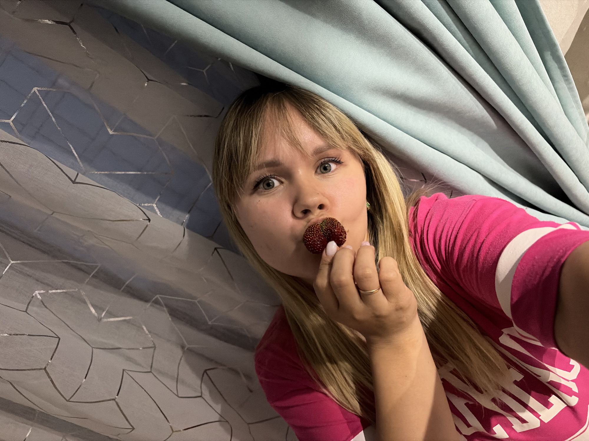 A blonde woman with fair skin, green eyes, and bangs is pictured holding a strawberry to her lips. She is wearing a bright pink t-shirt with white lettering that appears to spell out "PINK" in large block letters. The background consists of draped curtains, one a pale blue and the other a sheer white with a geometric silver pattern. The woman is positioned in the center of the frame, looking directly at the camera with a playful expression. Her hand, with a delicate ring on her finger, is visible near her mouth, holding the strawberry. The lighting suggests it is daytime, possibly indoors given the presence of curtains. The overall mood is lighthearted and casual. The image appears to be a selfie, taken from a slightly elevated angle. The woman's hair is long and blonde, falling over her shoulders.
