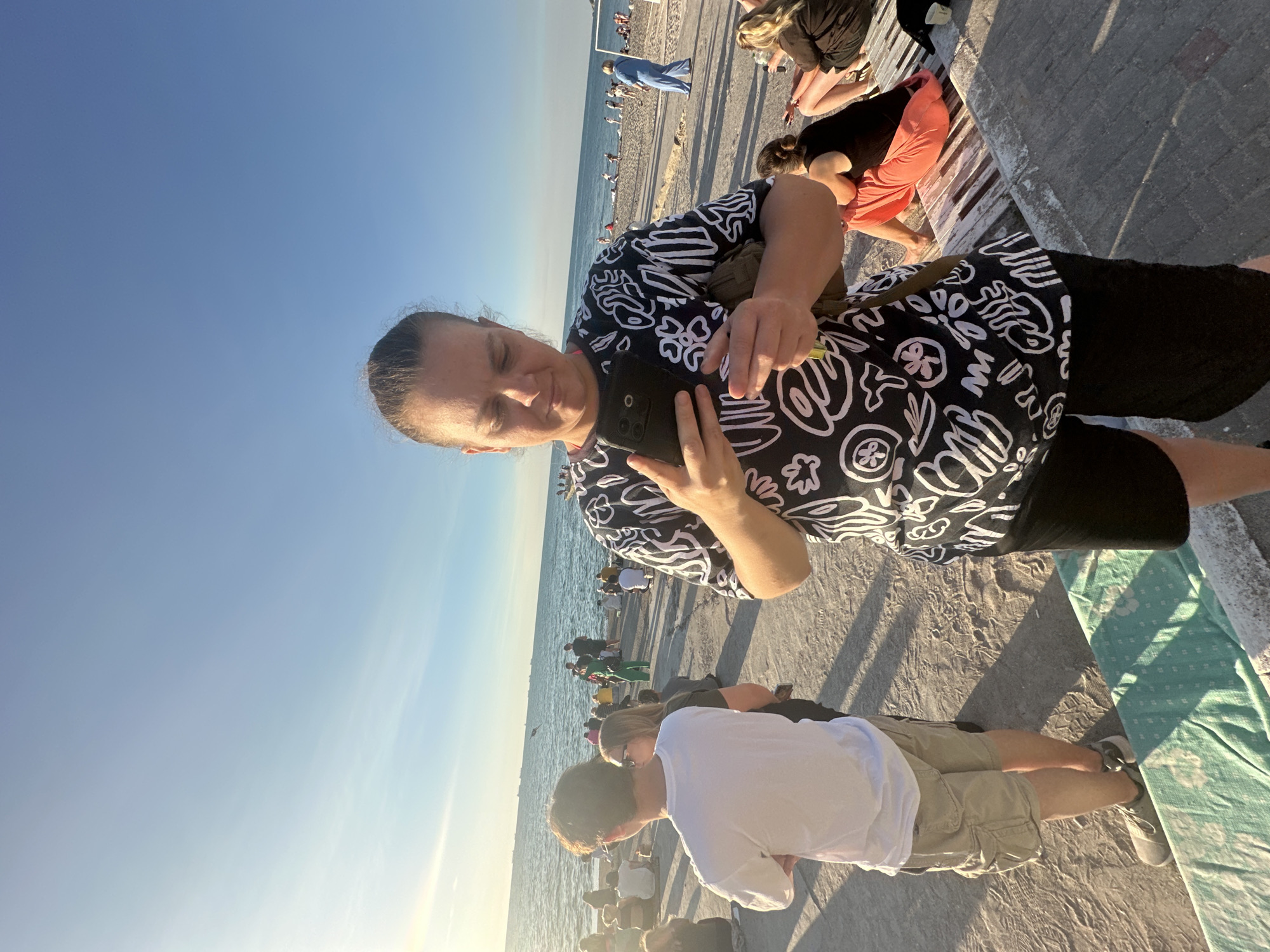 A woman in a black and white patterned shirt and black shorts stands on a concrete promenade, looking down at her smartphone, which she holds in both hands. She is in the foreground, with a beach and the sea visible behind her. People are gathered on the beach and along the promenade, some sitting on benches. The sun is shining, casting long shadows. The location is the Odesa seashore in Ukraine.