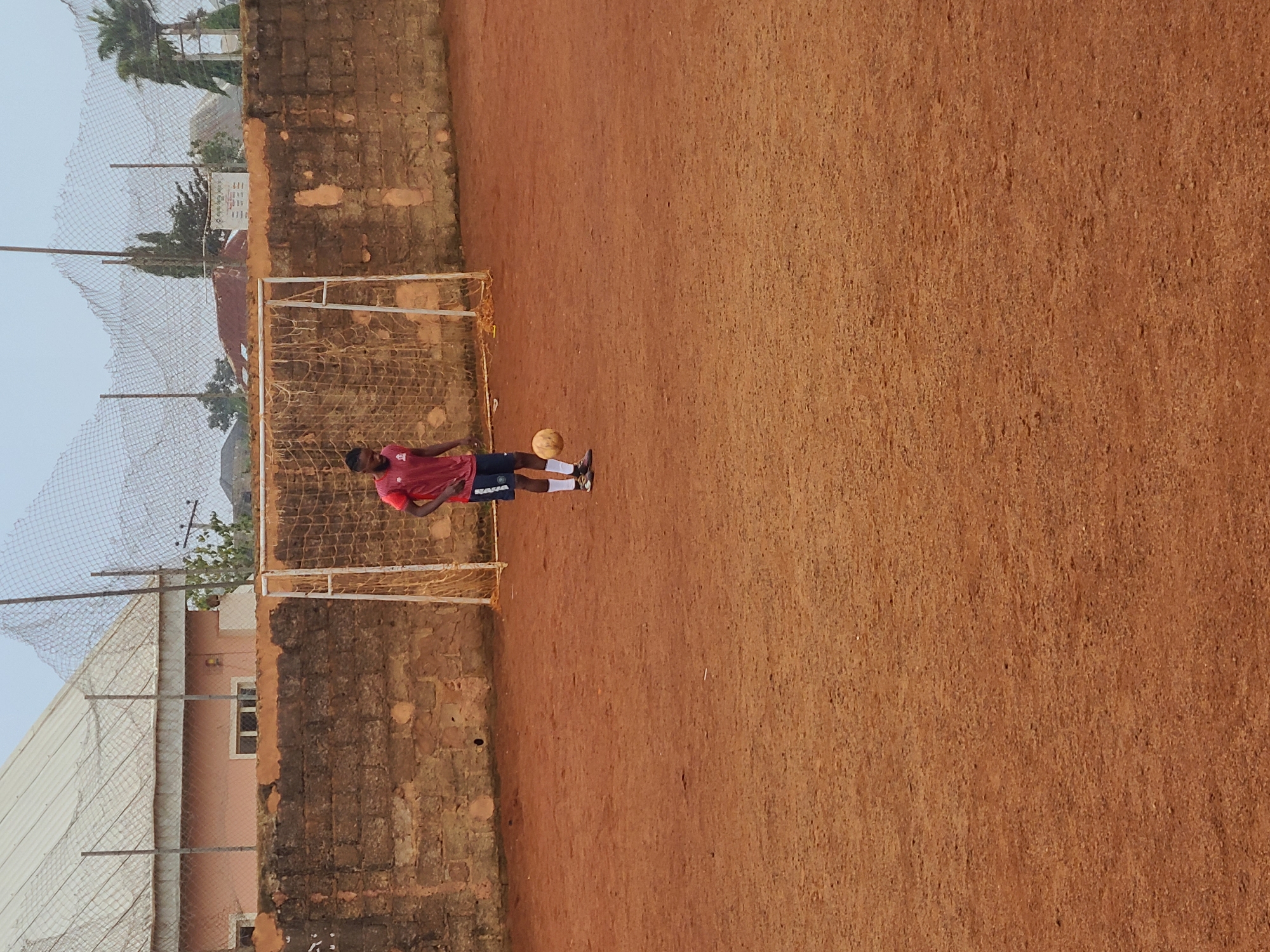 The image shows a person in a red shirt and dark shorts playing soccer on a dirt field. They are wearing white socks and cleats. A soccer ball is near their feet. In the background, there is a soccer goal with a net. Behind the goal, there is a brick wall, and beyond that, some buildings and trees. A fence surrounds part of the field. The sky is overcast, suggesting it might be early morning or late afternoon, or a cloudy day. The location is identified as Barakin Lerawi, Nigeria. There is some text visible on the person's shorts, which appears to say "NAHA".