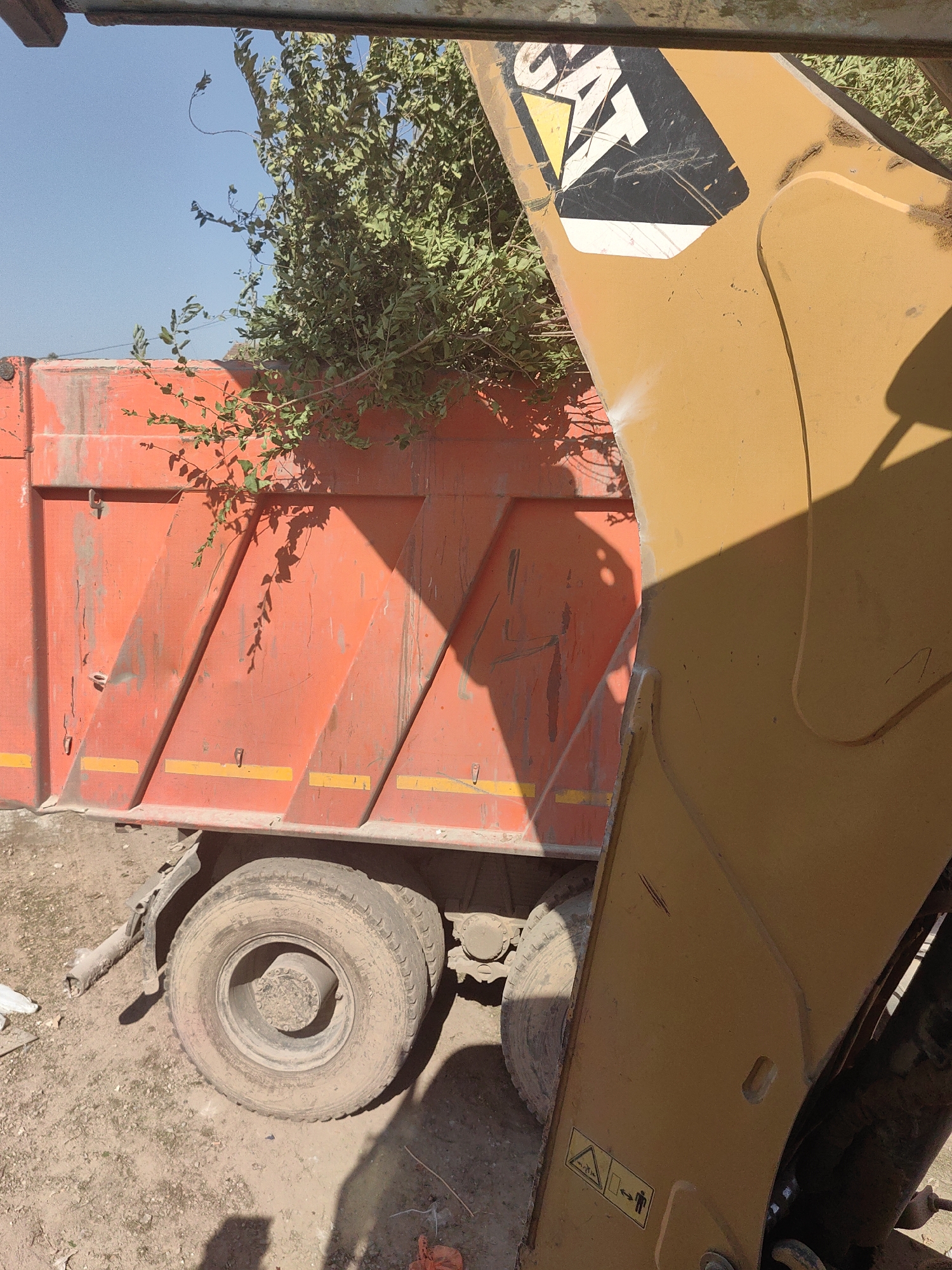 The image depicts a construction scene in Shymkent, Kazakhstan. A large orange dump truck is parked on a dirt surface. The truck bed is filled with green branches and foliage, suggesting debris removal or landscaping work. To the right of the truck, a yellow excavator arm is visible, bearing the "CAT" logo. The excavator is partially obscuring the truck. The sky is clear and blue. The ground is covered in loose soil and some scattered debris. The scene appears to be outdoors, possibly at a construction site or a public works area within the city.