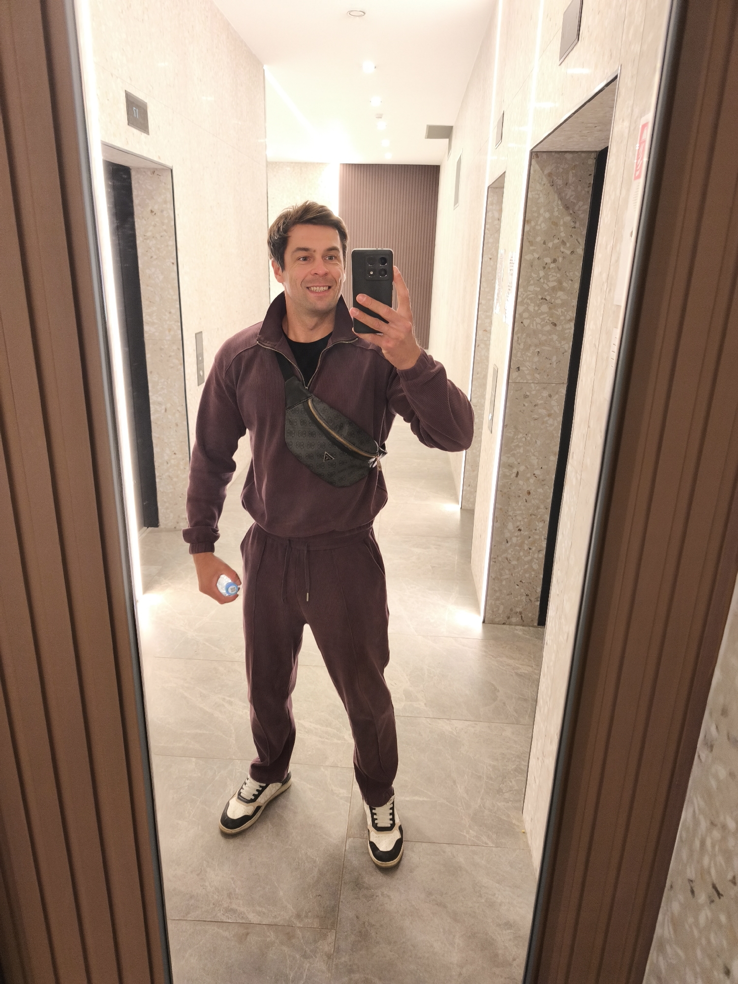 A man stands in a corridor, visible in a mirror reflection. He is holding a smartphone in his right hand, seemingly taking a selfie. He is wearing a purple tracksuit, a black T-shirt underneath, a black crossbody bag, and black and white sneakers. In his left hand, he is holding a water bottle.
The corridor features several elevator doors on both sides, with speckled wall coverings. The floor is tiled with a grey marble pattern. Overhead, there are recessed lights. The scene suggests a modern building interior.
Based on the interior design and the architecture, it is likely that the location is in Moscow, Russia.