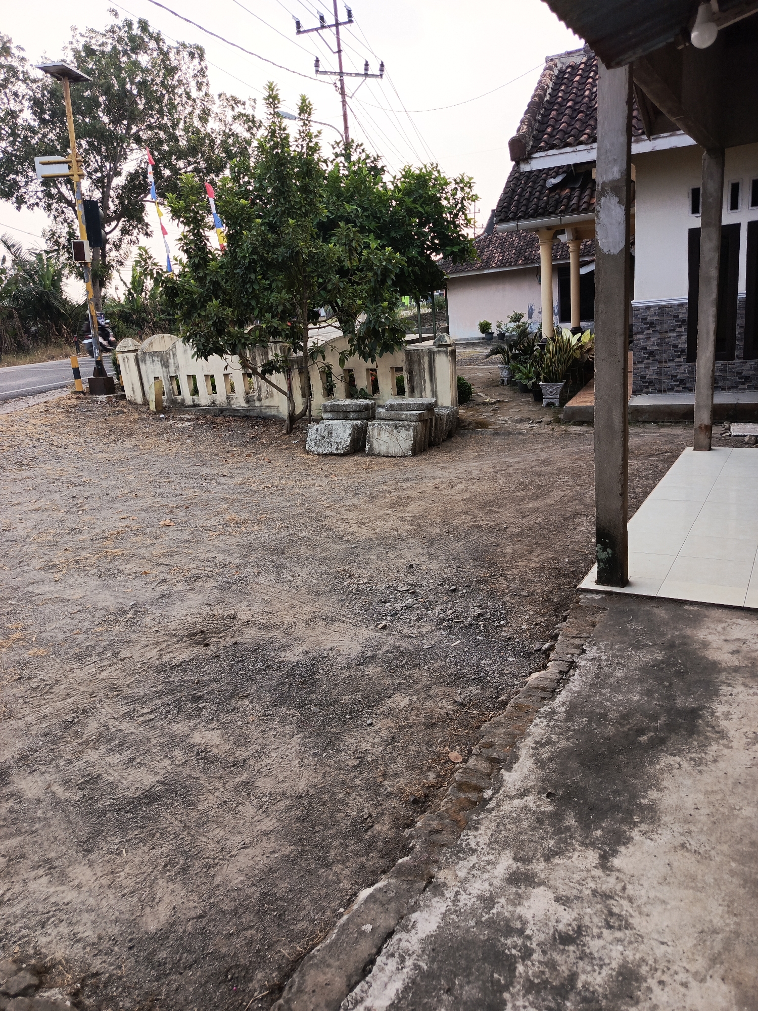 In the village of Karangsari, Indonesia, an exterior shot captures a roadside scene with a house on the right. The foreground is a dirt and gravel pathway. To the left, a road runs parallel to a low, decorative concrete wall with rectangular openings. Behind the wall, a tree with dense green foliage stands. A traffic light pole with a solar panel is visible near the road. In the background, a house with a tiled roof and white walls is partially visible, with several potted plants in front. Electrical wires crisscross the sky. The overall impression is of a rural Indonesian setting.