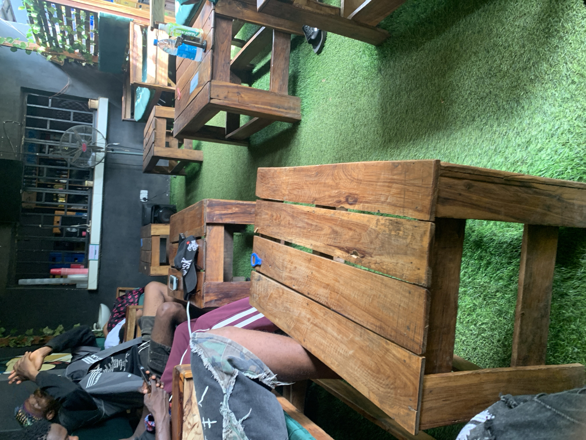 The image shows an outdoor seating area with several wooden tables and chairs placed on artificial turf. There are at least three individuals visible, with one person in the foreground sitting on a wooden bench with their legs crossed, wearing ripped denim shorts and a maroon track pant. Another person, seen from the waist up, is seated at a table, holding a phone and wearing a black jacket. A third person, partially visible on the left, appears to be wearing a patterned headwrap and a black jacket. In the background, a bar area with shelves stocked with bottles and a fan is visible. The lighting suggests it is daytime, with sunlight casting shadows on the ground. The setting appears to be a casual outdoor gathering or a small eatery.