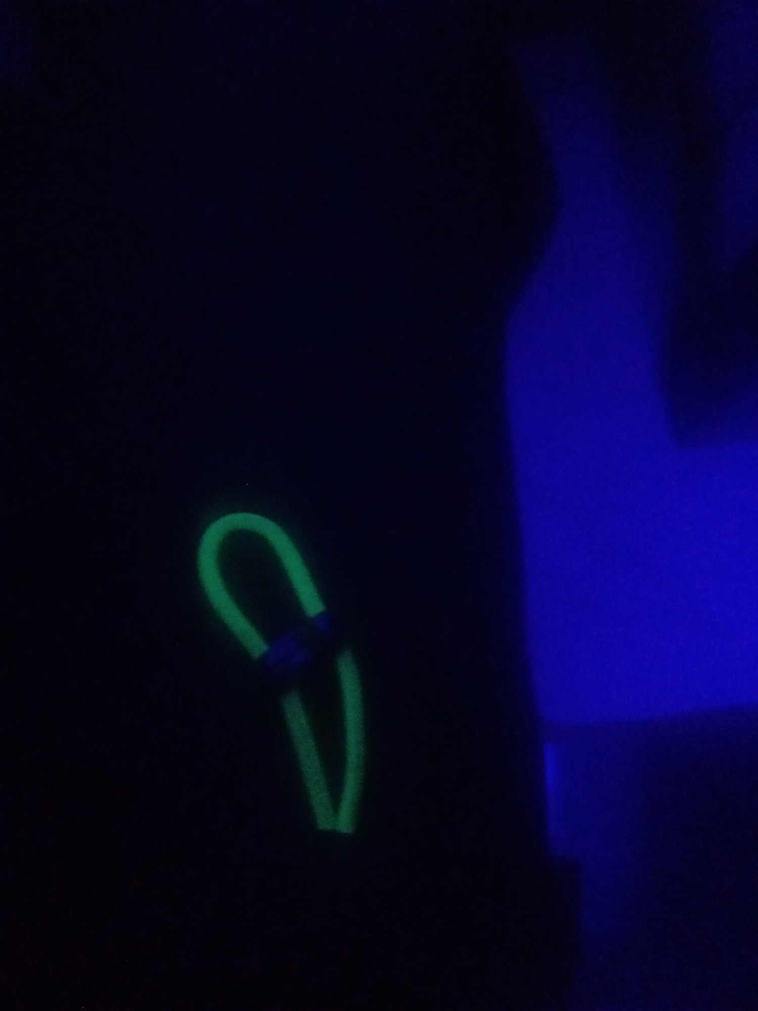 In a dimly lit environment, a neon green loop is visible, tied with a dark blue string. The loop, which appears to be made of rope or cord, is positioned in the lower left quadrant of the image. The background is predominantly black, with a soft, electric blue glow emanating from the right side of the frame. The blue light casts an ethereal sheen on the dark surroundings, suggesting an artificial light source. No people or other objects are discernible in the scene. The location is identified as Unknown, Unknown.