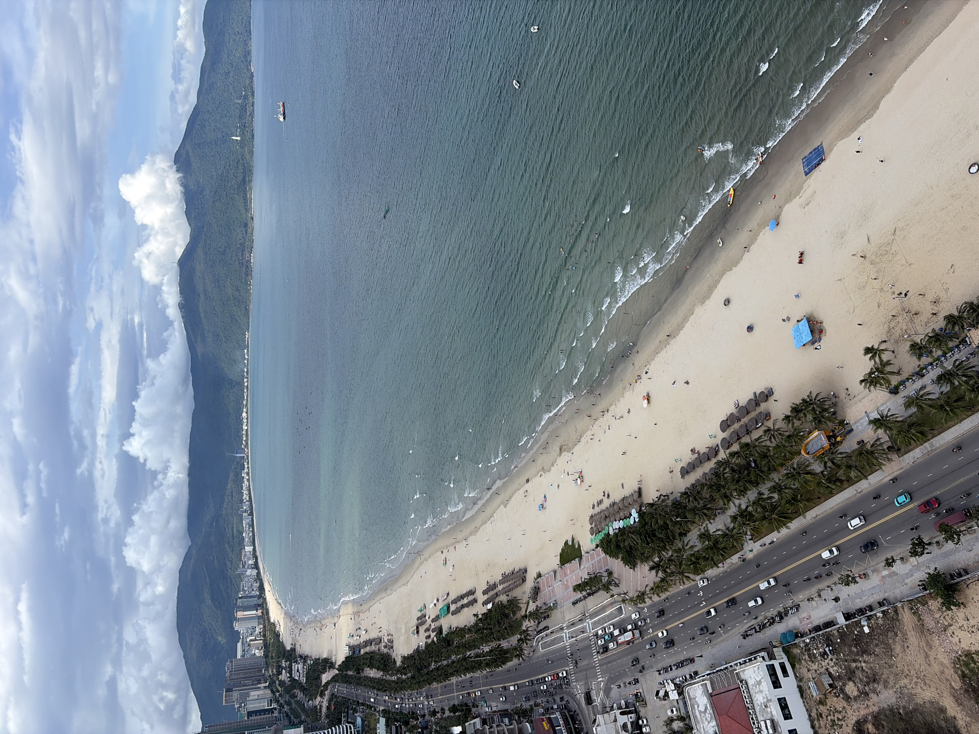 This is an aerial, high-angle shot of a coastal area in Da Nang, Vietnam. The scene depicts a wide, sandy beach with numerous sunbathers, umbrellas, and thatched-roof loungers. The ocean is a deep teal color with moderate waves breaking on the shore. A busy coastal road runs parallel to the beach, with a continuous flow of cars and motorbikes. Palm trees line the sidewalk between the road and the beach. In the background, a city skyline with tall buildings stretches along the coast, backed by a range of green, tree-covered mountains under a cloudy sky. Several boats are visible in the ocean.