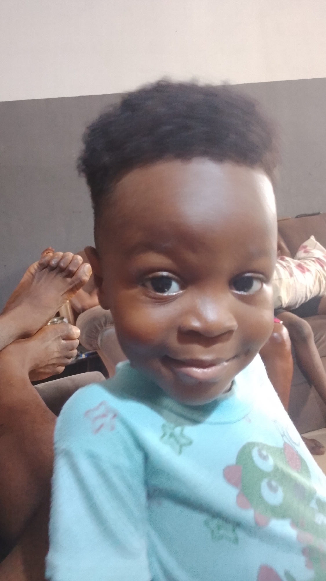 A young child with dark skin, close-cropped dark hair, and wearing a light blue shirt with a cartoon design is the central subject of the image. The child is smiling slightly and looking directly at the camera. In the background, out of focus, are human feet and legs resting on what appears to be a brown upholstered couch. The wall behind is two-toned, with grey on the bottom and off-white on top. Given the request for a location of Lokoja, Nigeria, this scene likely represents a casual domestic setting there.