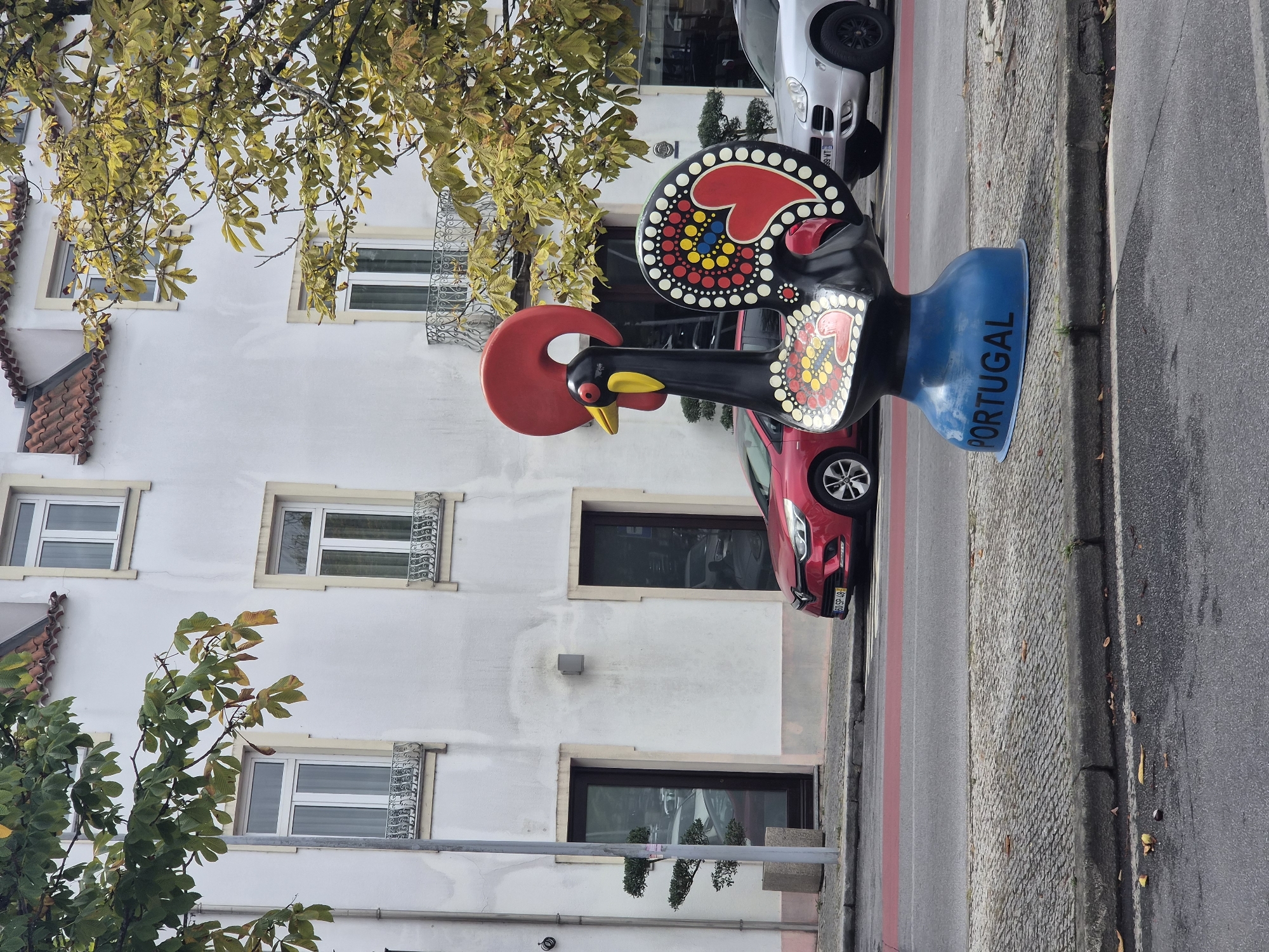 The image shows a Barcelos Rooster statue, a symbol of Portugal, standing on a sidewalk. The statue is black with red and yellow accents and has "PORTUGAL" written on its blue base. Behind the statue, a white building with red tile roofs and multiple windows is visible. A red car is parked in front of the building, and a silver SUV is to the right of the statue. Trees with yellowing leaves are also present in the scene, suggesting an autumn setting. The statue is located in the city of Barcelos, Portugal, a place renowned for its pottery and the origin of the Barcelos Rooster legend.